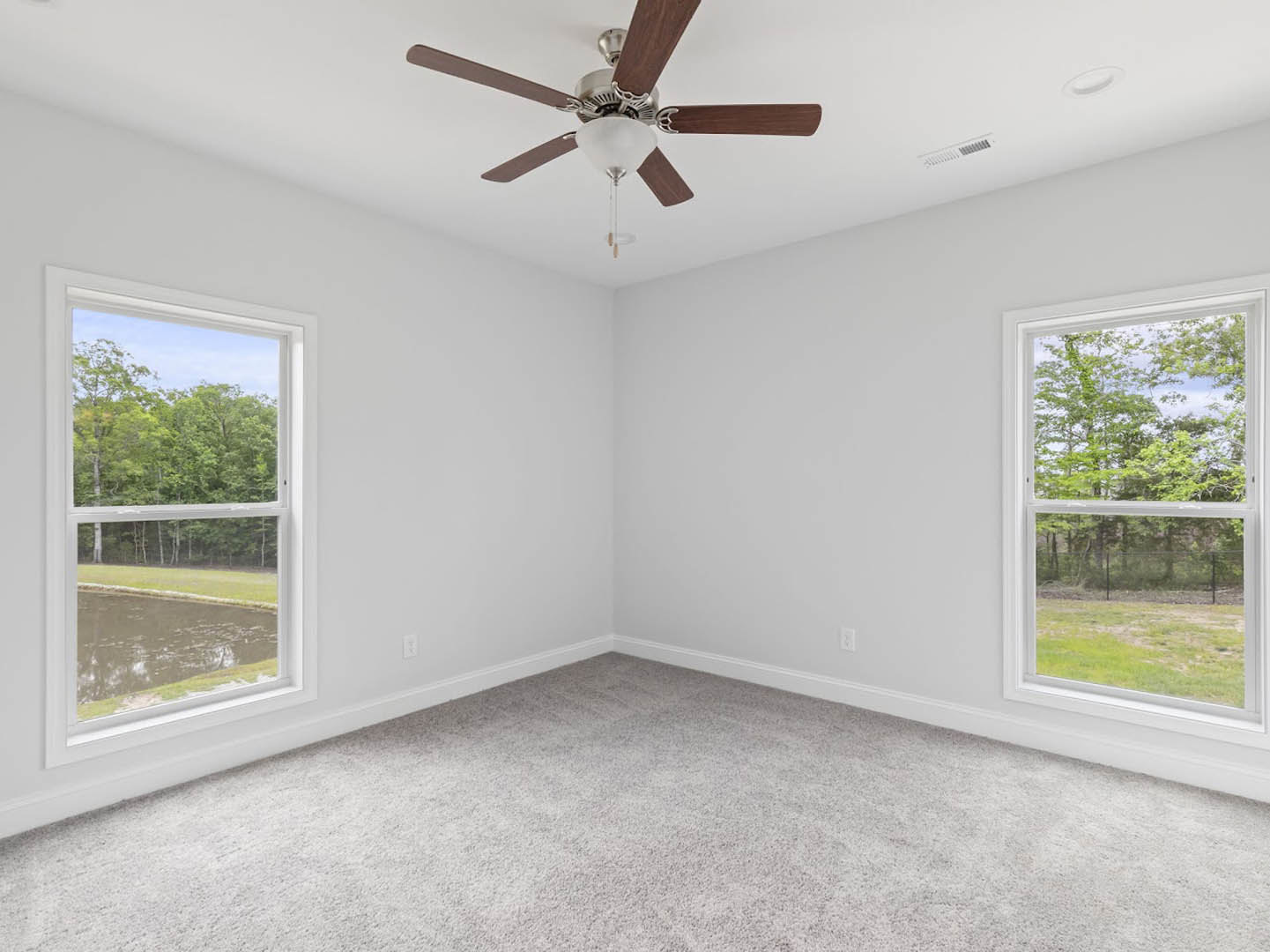 Carpeted room with white plaster walls, ceiling fan with light fixture, large windows overlooking trees and water.