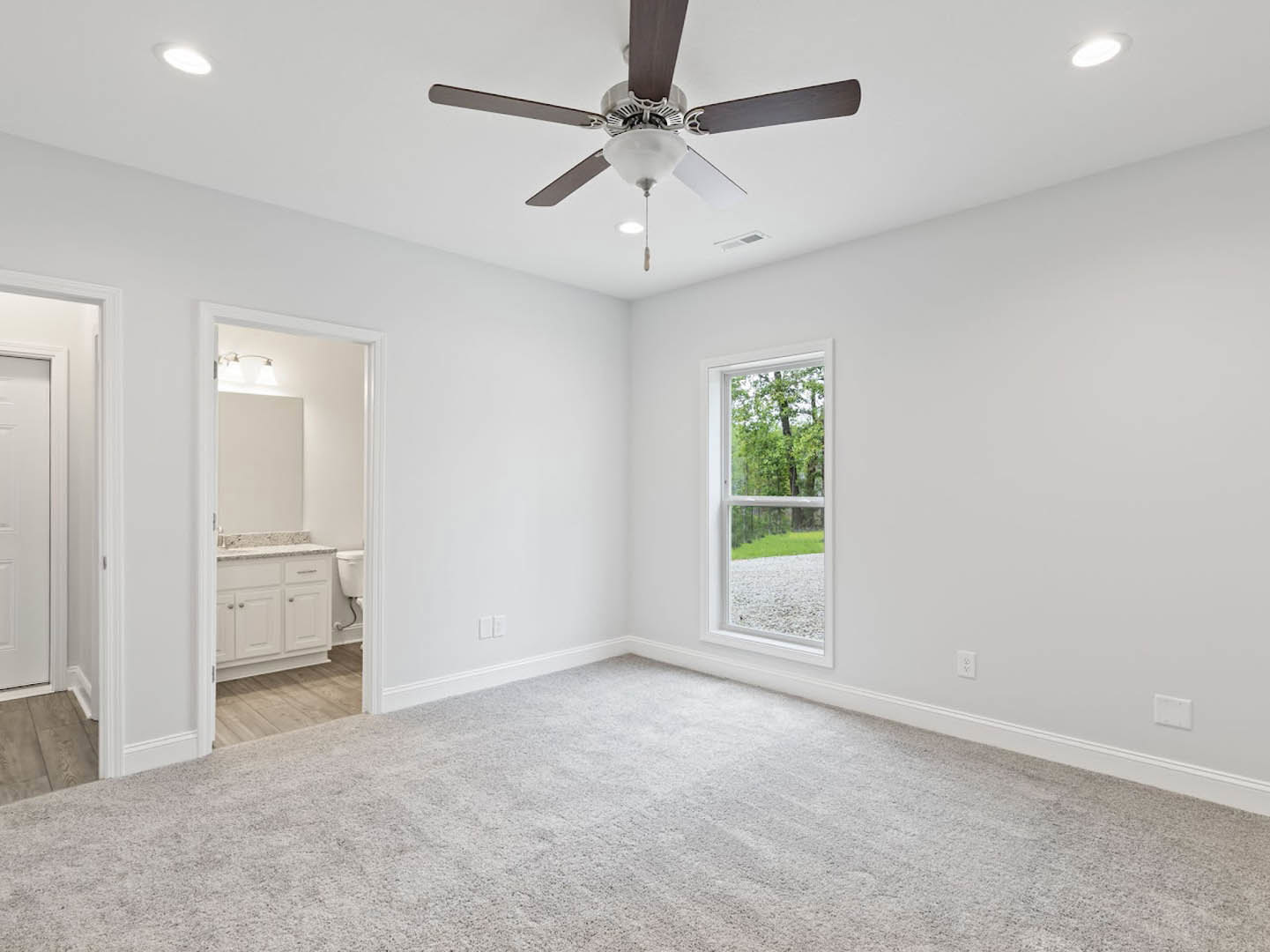 Bedroom with white carpet, ceiling fan with light fixture, large window showing trees outside, adjacent bathroom featuring sink and toilet