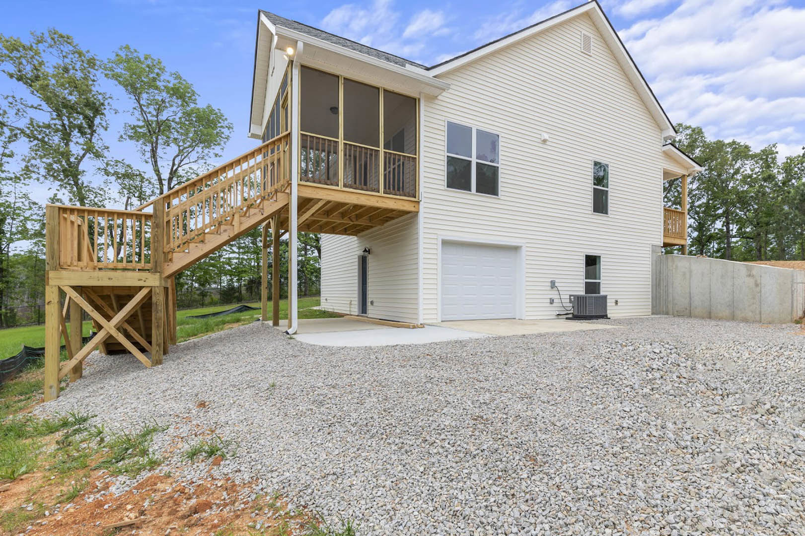 Two-story home with white siding, attached garage featuring a white door, elevated wooden deck, gravel driveway, large windows, and surrounding trees under a cloudy sky
