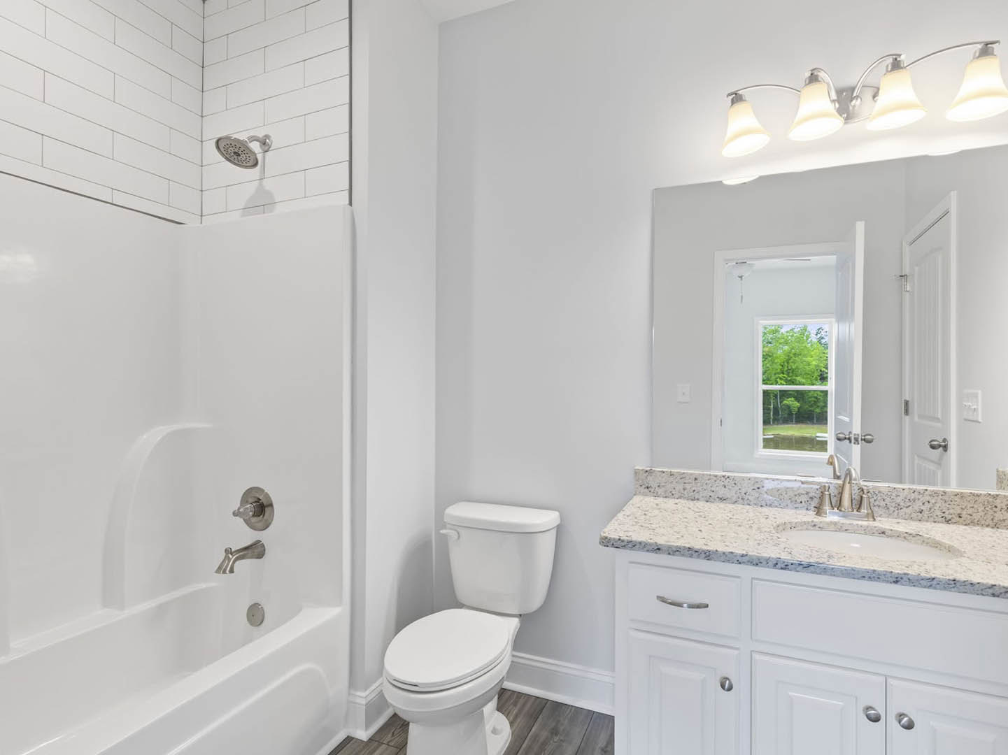Bathroom with white toilet, open lid, rectangular sink on vanity, wall-mounted mirror, two-bulb light fixture, tiled walls, shower head, window showing green trees outside