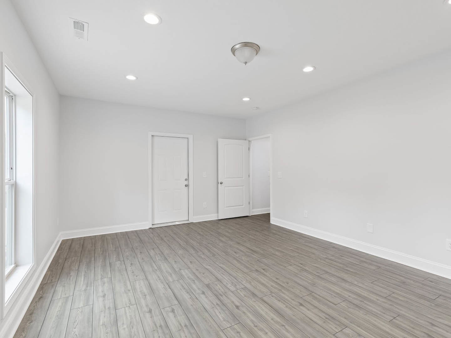 White walls, wood flooring, white door with silver knobs, ceiling light fixture, window on white wall