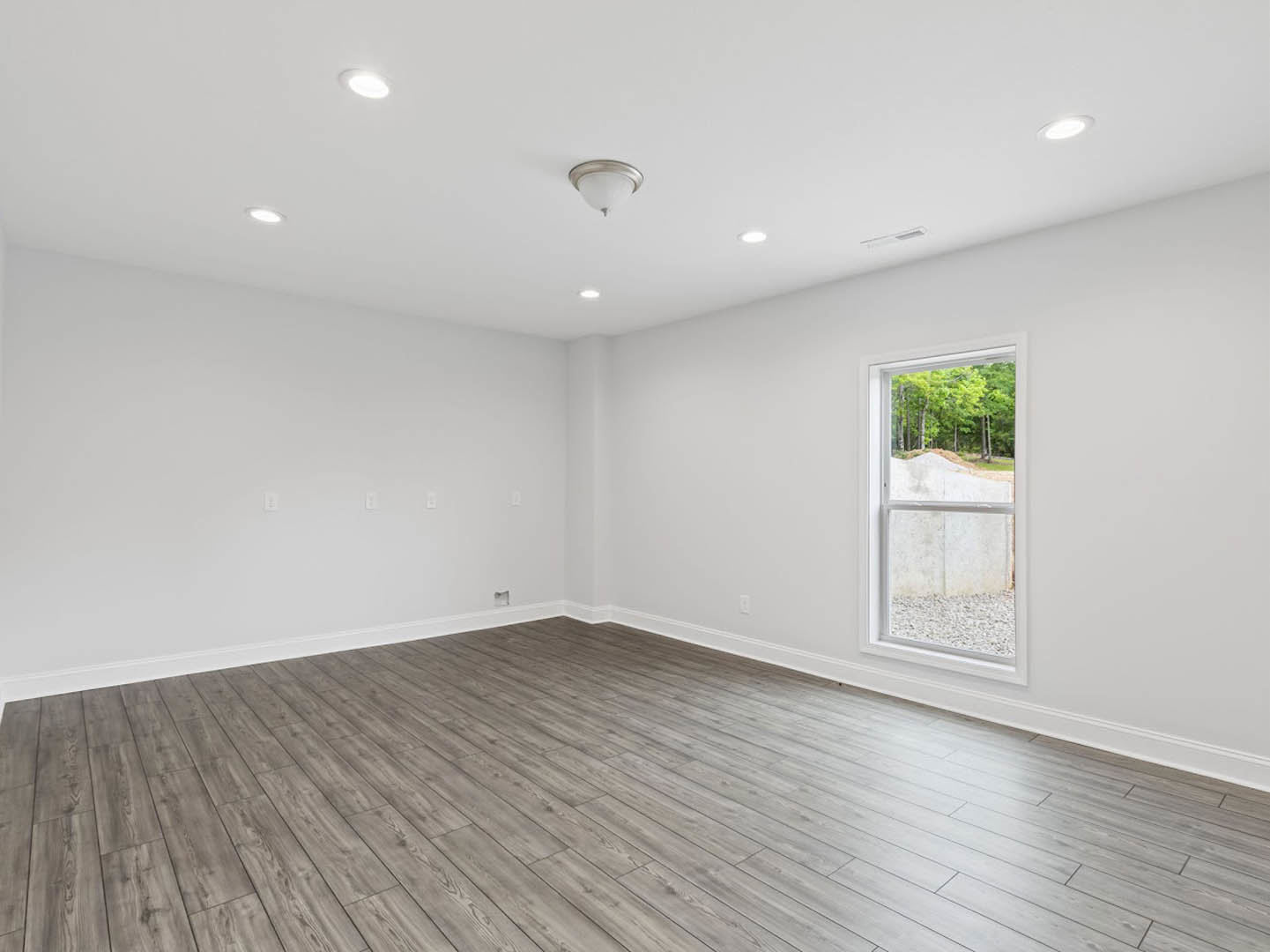 Sunlit room with wide window overlooking forest, light wood flooring, white plaster walls, and modern ceiling fixture