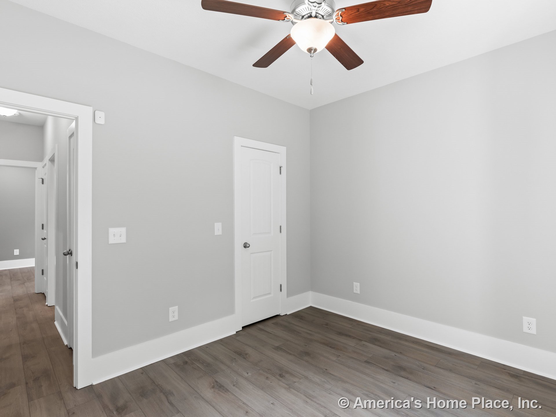 Bedroom with light gray walls, wide white baseboards, wood-look plank flooring, white paneled door, wood ceiling fan with light, and recessed hallway lighting.