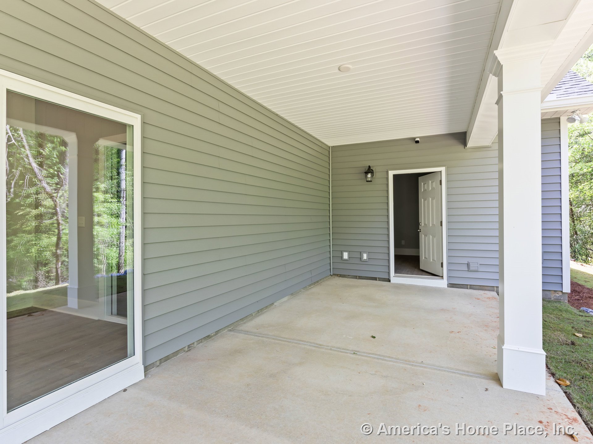 Covered porch with gray horizontal siding, large sliding window, white paneled ceiling, concrete floor, white trim and columns, and exterior wall-mounted light fixture.