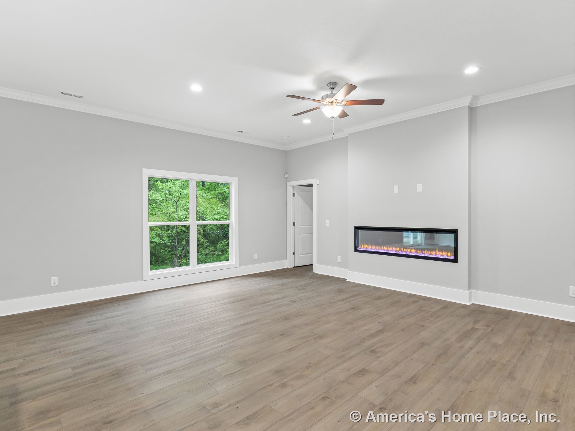 Modern living room with linear wall fireplace, wide plank wood flooring, large double window with views of greenery, ceiling fan with light, recessed ceiling lights, white trim and