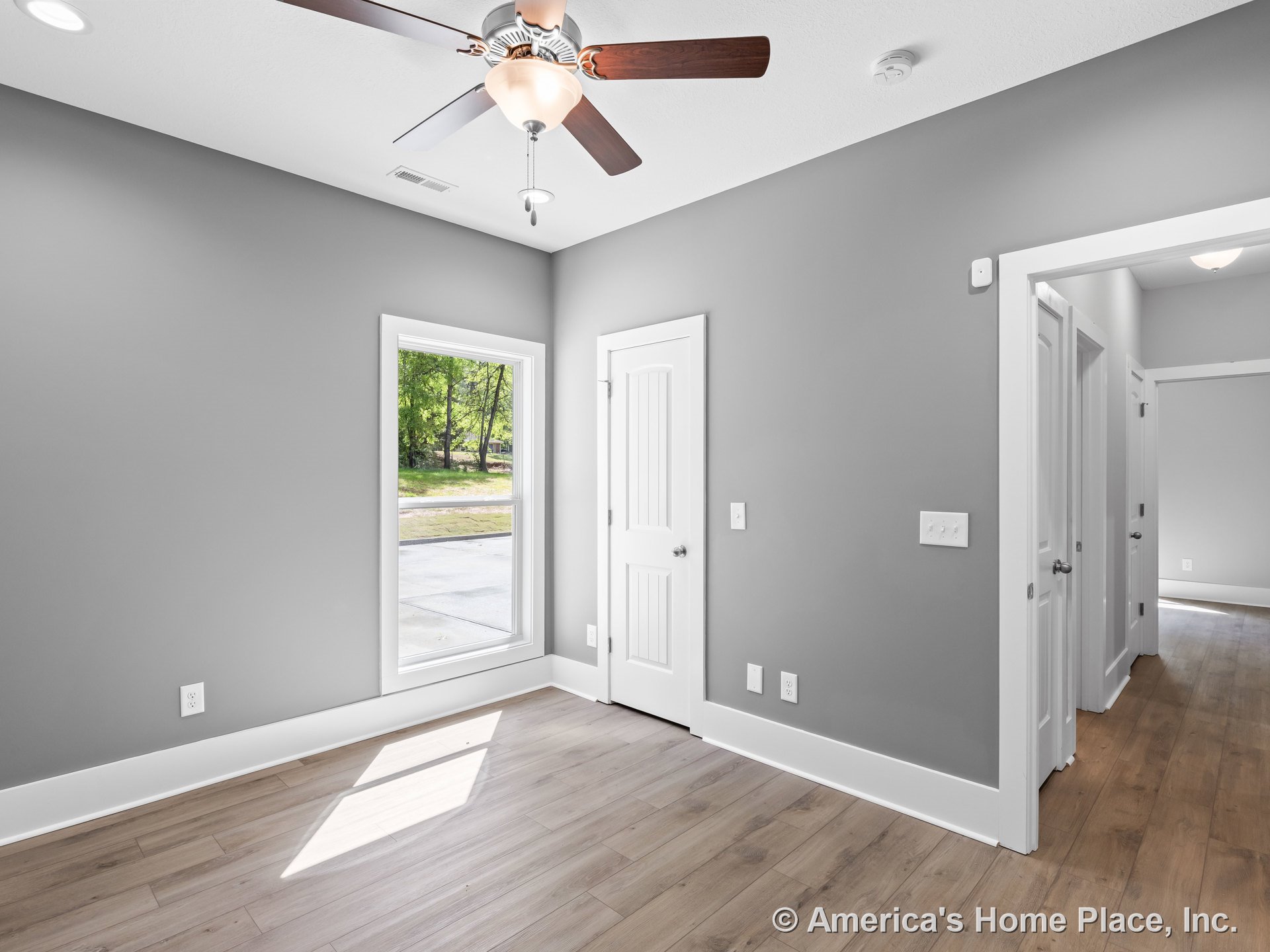 Bedroom with gray painted walls, wide white baseboards, large window with white casing, paneled white doors, wood-look plank flooring, and ceiling fan with integrated light