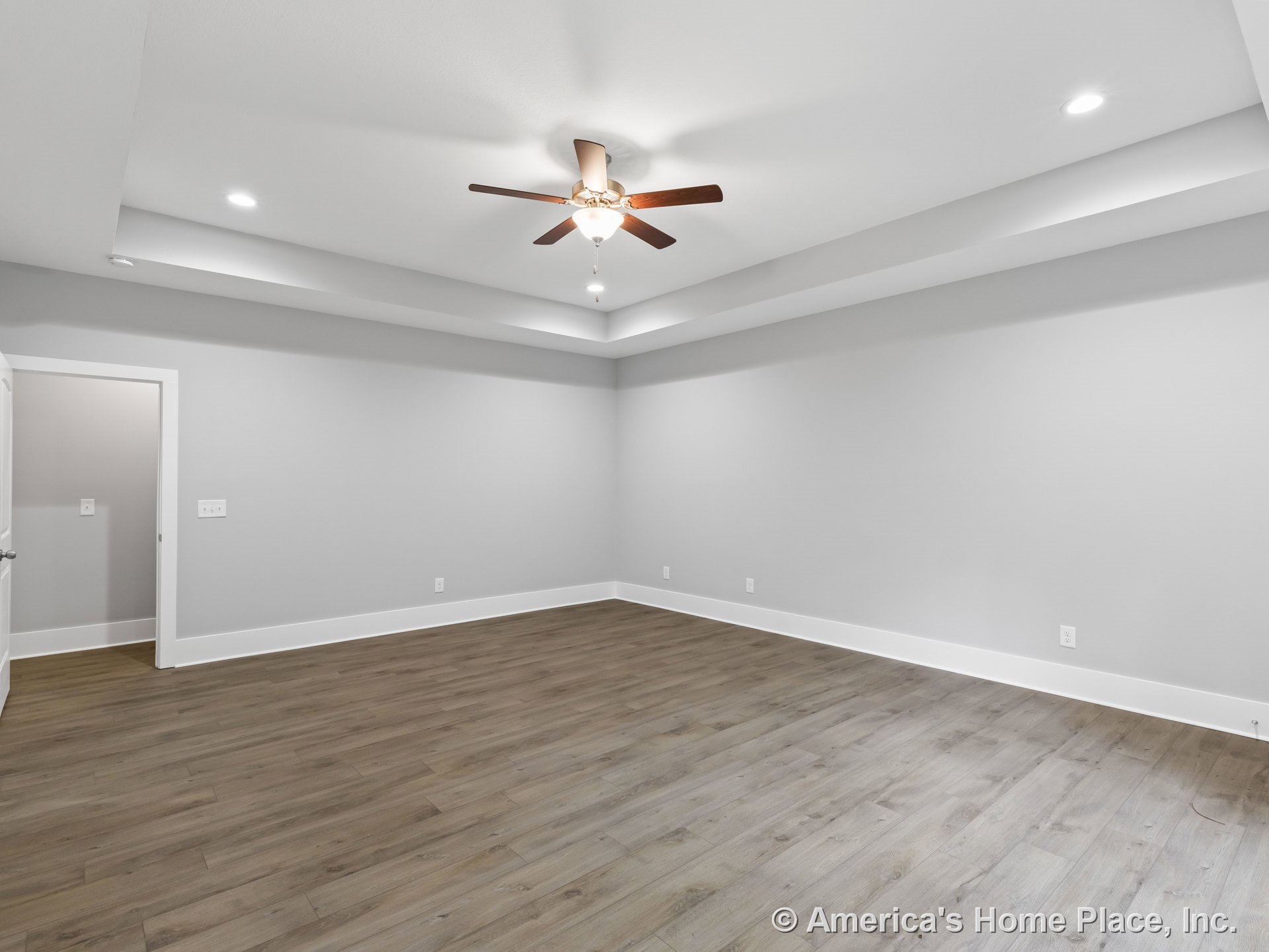 Tray ceiling with recessed lights and ceiling fan, wide baseboards, wood-look plank flooring, neutral gray walls, white trim surrounding door, spacious empty room with modern