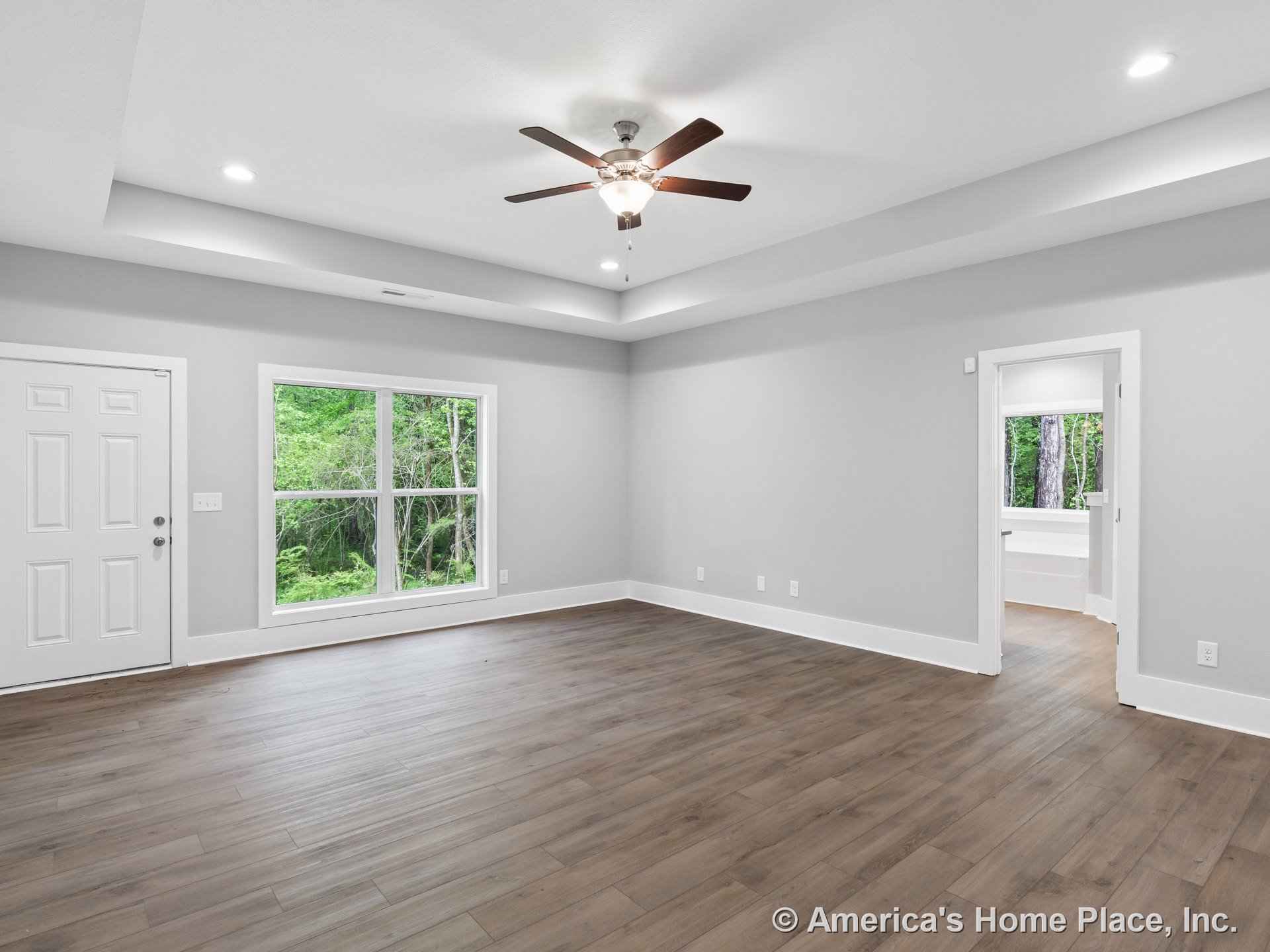 Wide plank wood flooring, tray ceiling with recessed lighting and ceiling fan, large double window with white trim, entry door with panel design, and crisp white baseboards.