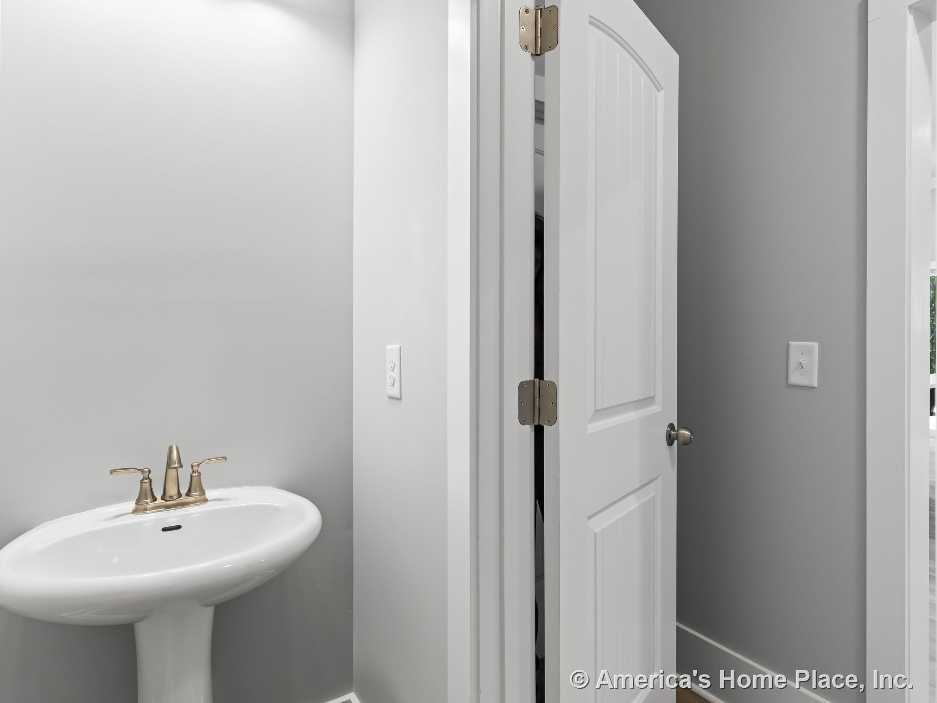 Pedestal sink with brushed nickel faucet, gray painted walls, white paneled door, white trim, recessed ceiling light, minimalist baseboard detailing.