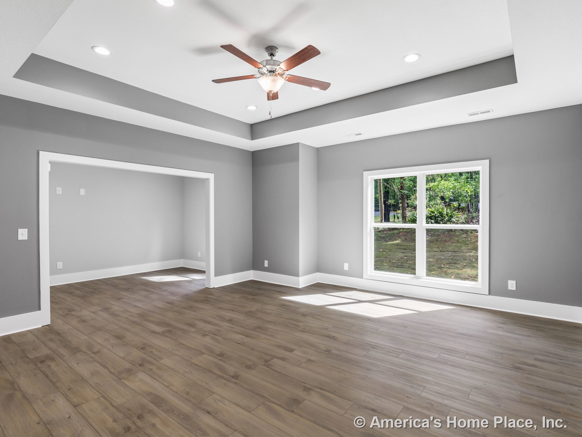 Tray ceiling with ceiling fan and recessed lighting, large double window allowing natural light, wood-look plank flooring, wide doorway with white trim, gray painted walls, open