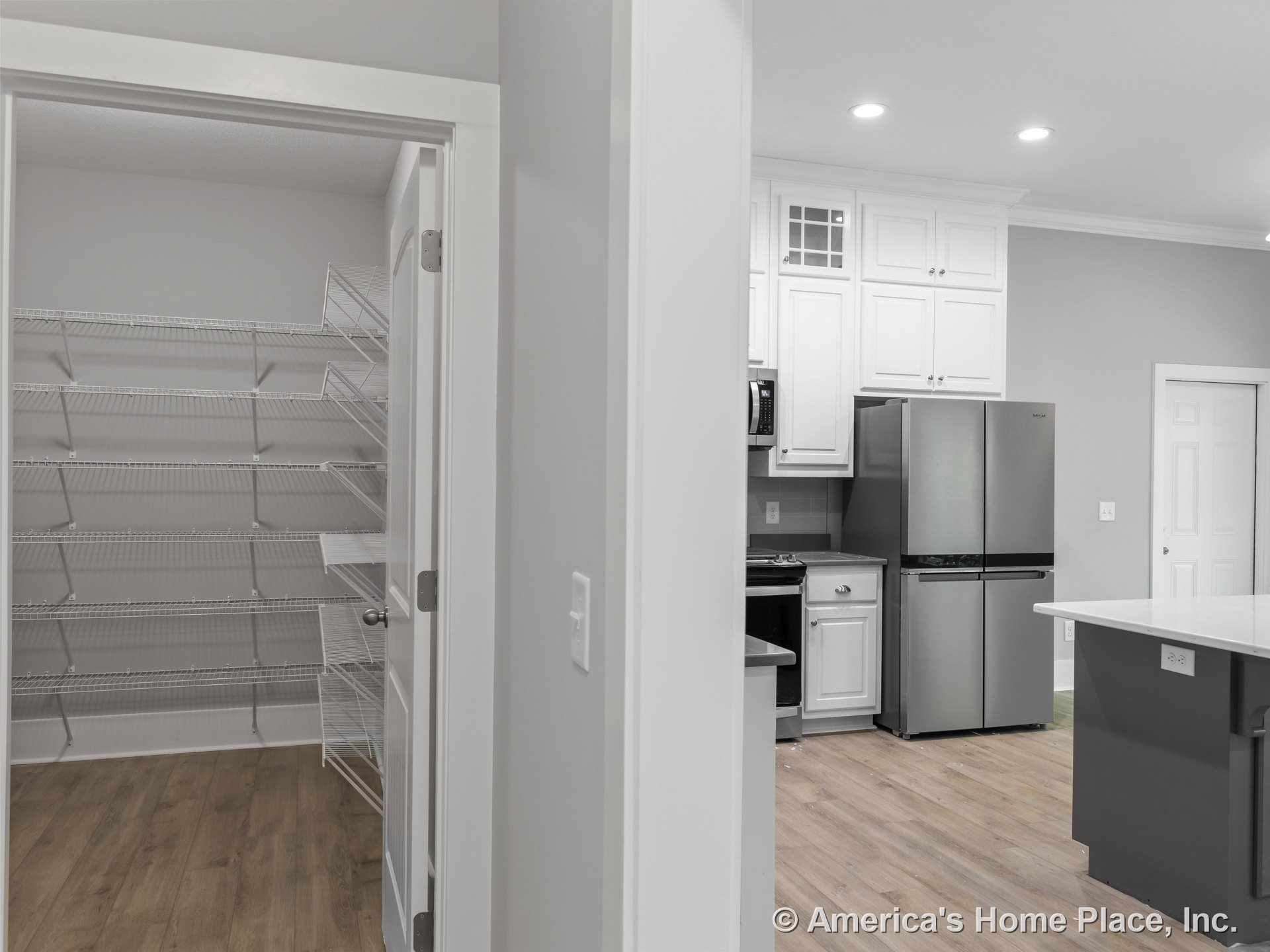 White upper and lower kitchen cabinets, stainless steel refrigerator, gray kitchen island with quartz countertop, recessed ceiling lights, light wood flooring, and walk-in pantry