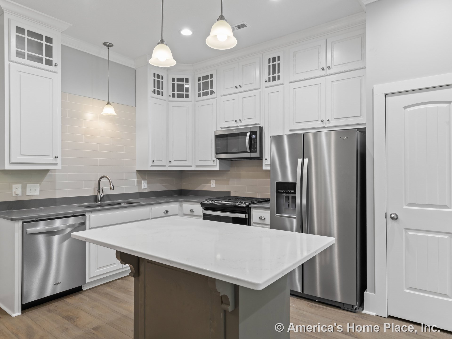 White kitchen with upper cabinets featuring glass inserts, subway tile backsplash, stainless steel appliances including refrigerator, built-in microwave above stove, central island