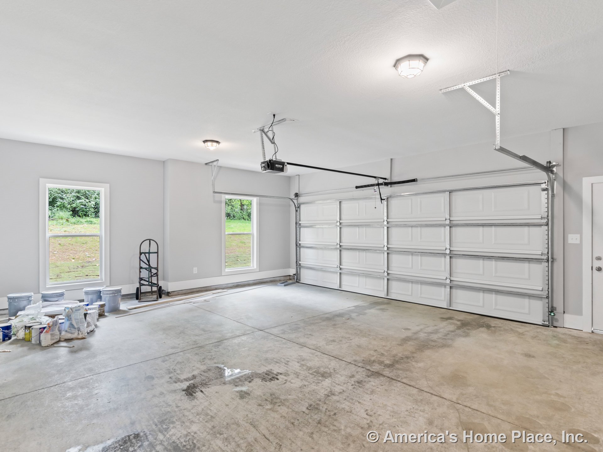 Double garage with paneled doors, concrete flooring, two windows for natural light, ceiling-mounted light fixtures, gray painted walls, and a side entry door with trim.