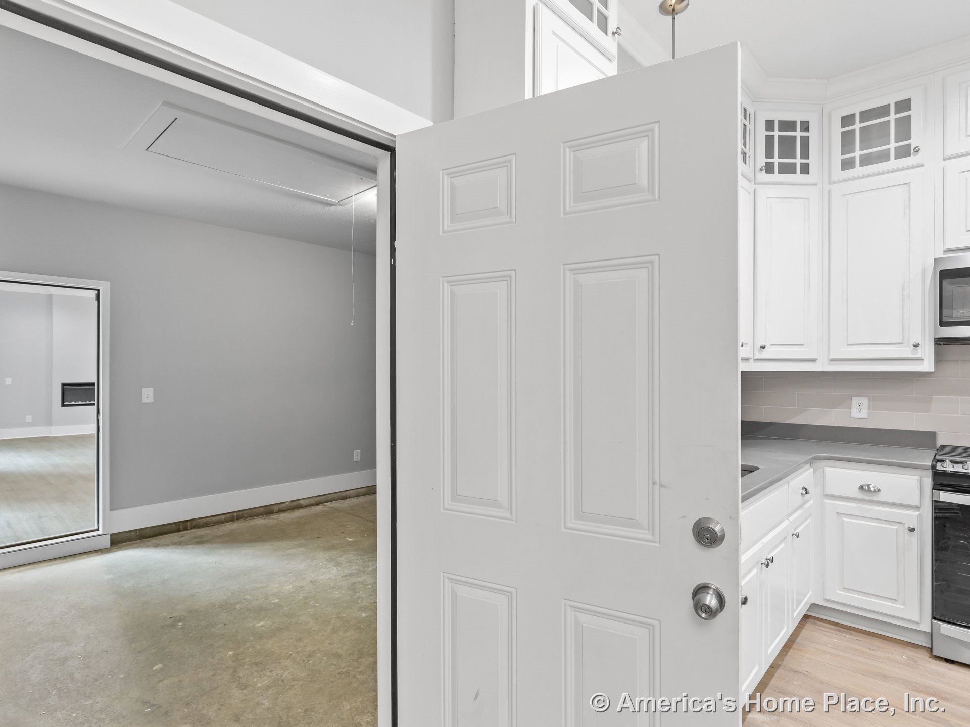White upper and base kitchen cabinets with gray solid surface countertops, stainless steel microwave, tile backsplash, built-in lighting, and adjacent garage featuring concrete