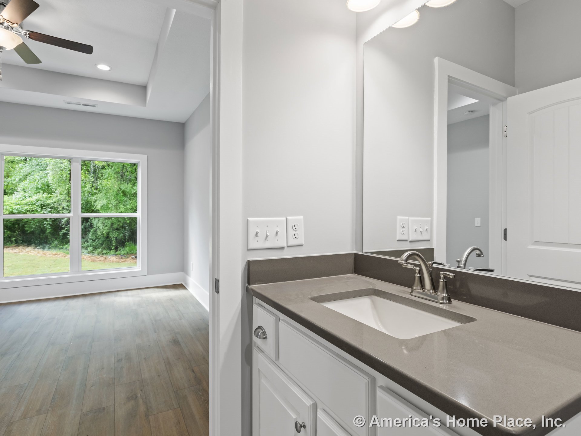 Bathroom vanity with gray solid surface countertop, undermount sink, brushed nickel faucet, and white cabinetry next to bedroom featuring large windows, wood-look flooring, and