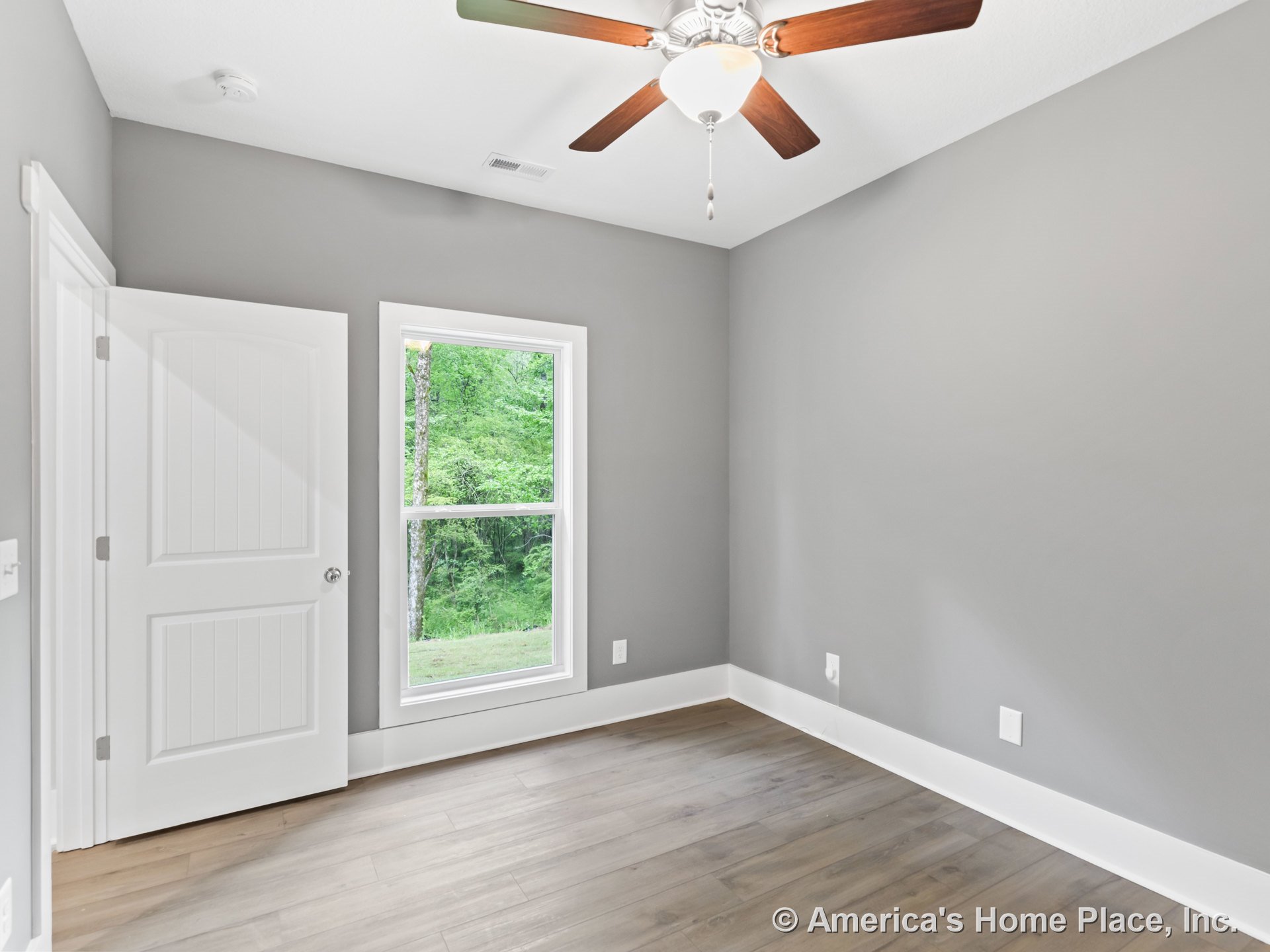Bedroom with gray walls, white baseboards and door trim, wood plank flooring, large window with white casing, ceiling fan and light fixture, paneled white door, modern finishes