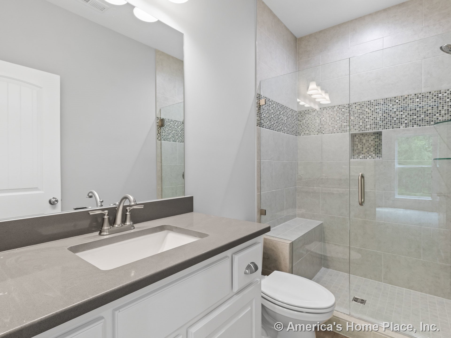 Gray solid surface countertop atop white shaker-style vanity cabinetry, frameless glass shower enclosure with mosaic tile accent strip, built-in shower bench, recessed niche in