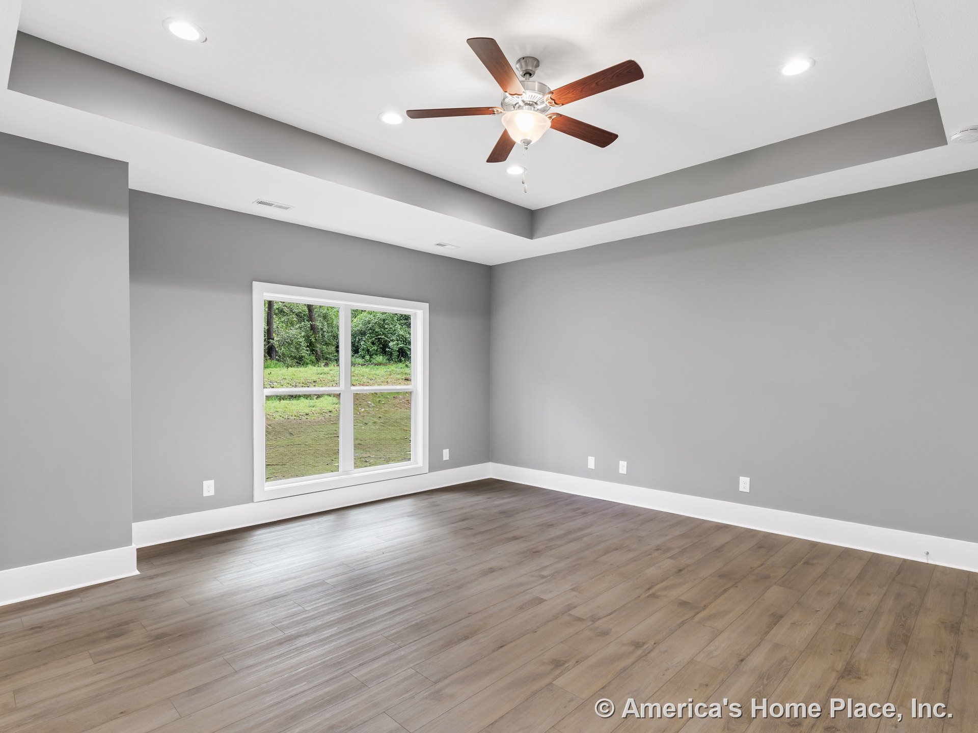 Tray ceiling with recessed lighting and ceiling fan, large window allowing natural light, wood-look flooring, gray walls with modern baseboards, empty room showcasing contemporary