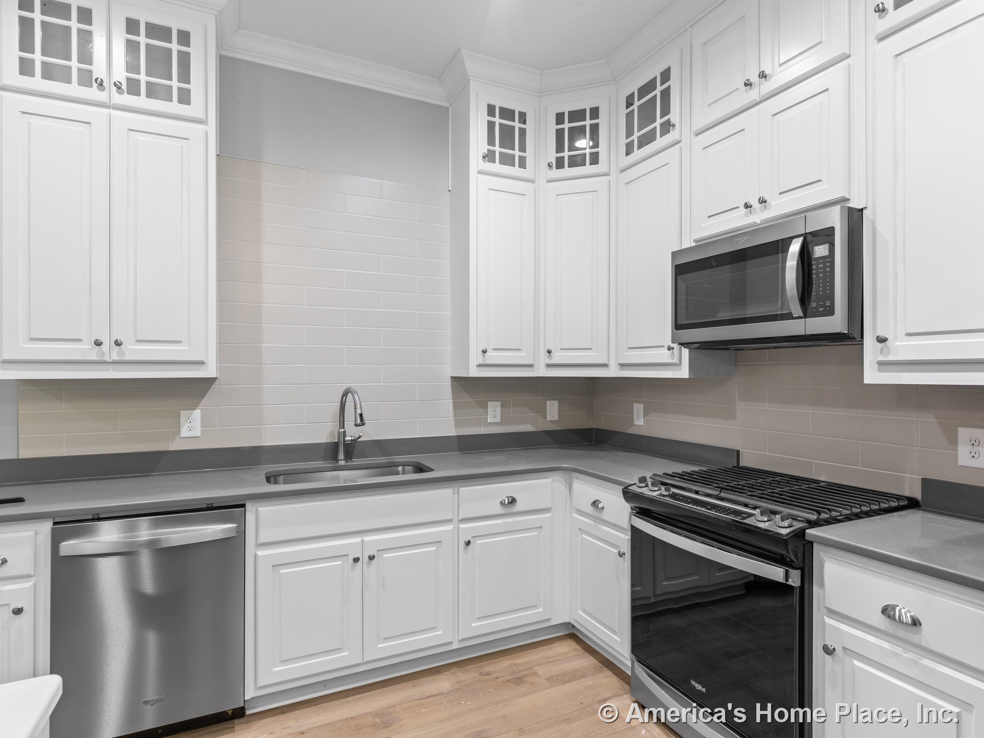 White kitchen with upper cabinets featuring glass inserts, gray countertops, subway tile backsplash, built-in stainless steel microwave, gas range, undermount sink, and wood