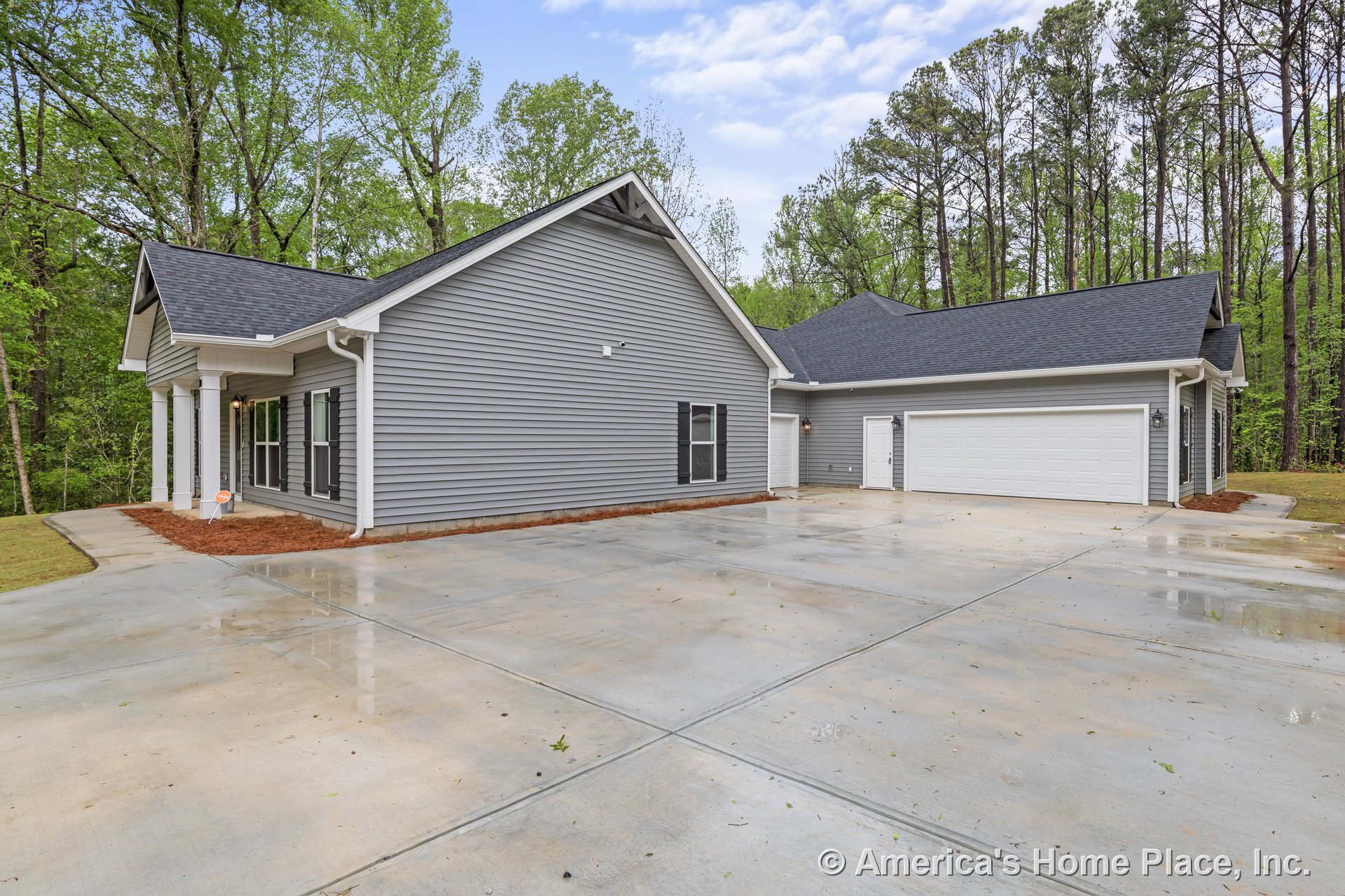 Gray vinyl siding exterior with white trim and gutters, covered front porch supported by columns, two-car garage with white doors, expansive concrete driveway, black roof shingles