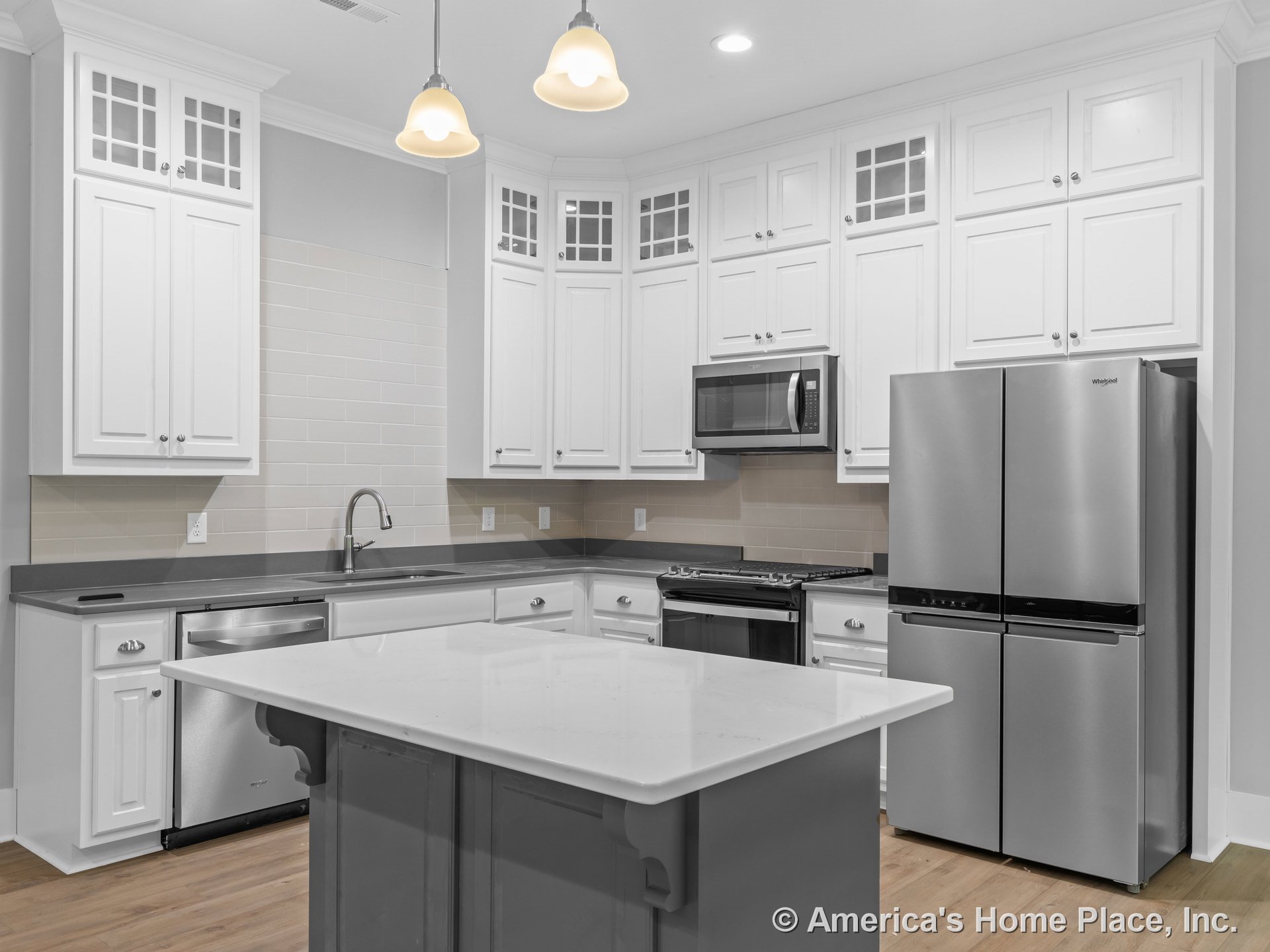 White shaker upper cabinets with glass inserts, stainless steel refrigerator and built-in microwave, subway tile backsplash, central island with quartz countertop, pendant ceiling