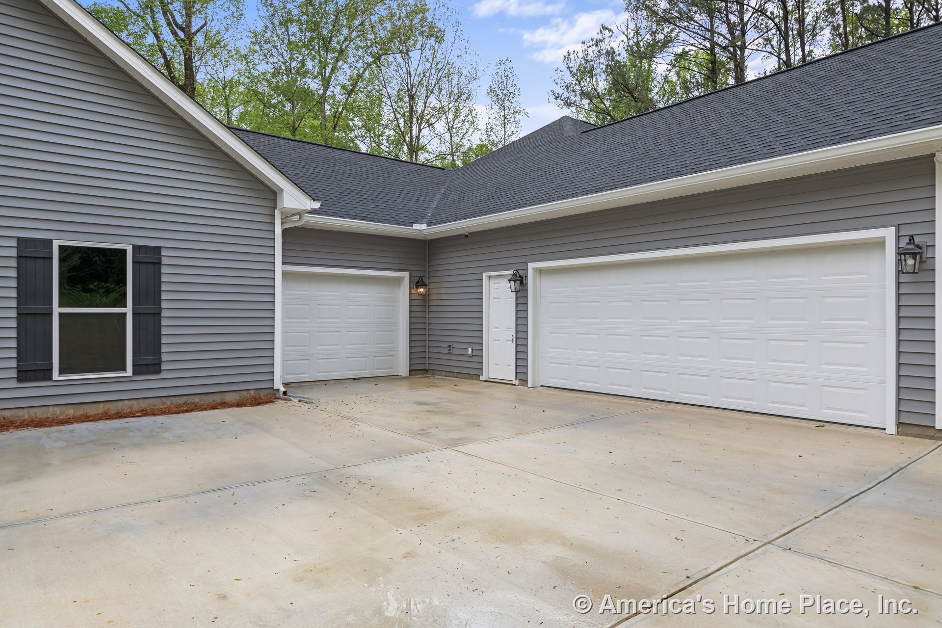 Double white garage doors set in gray vinyl siding with black window shutters, exterior wall lanterns, white trim and gutters, and a concrete driveway.