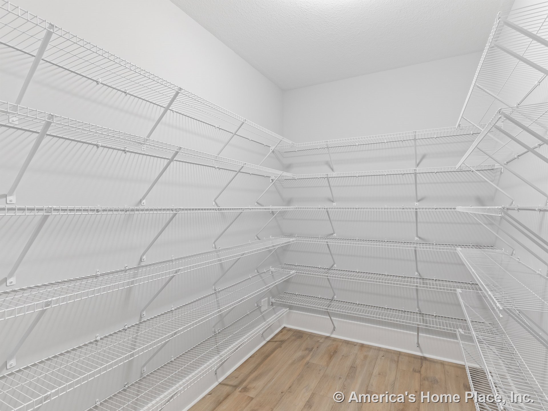 Walk-in pantry with multiple white wire shelving units, light wood flooring, white walls, flat ceiling, and built-in storage for organized food and kitchen supplies.