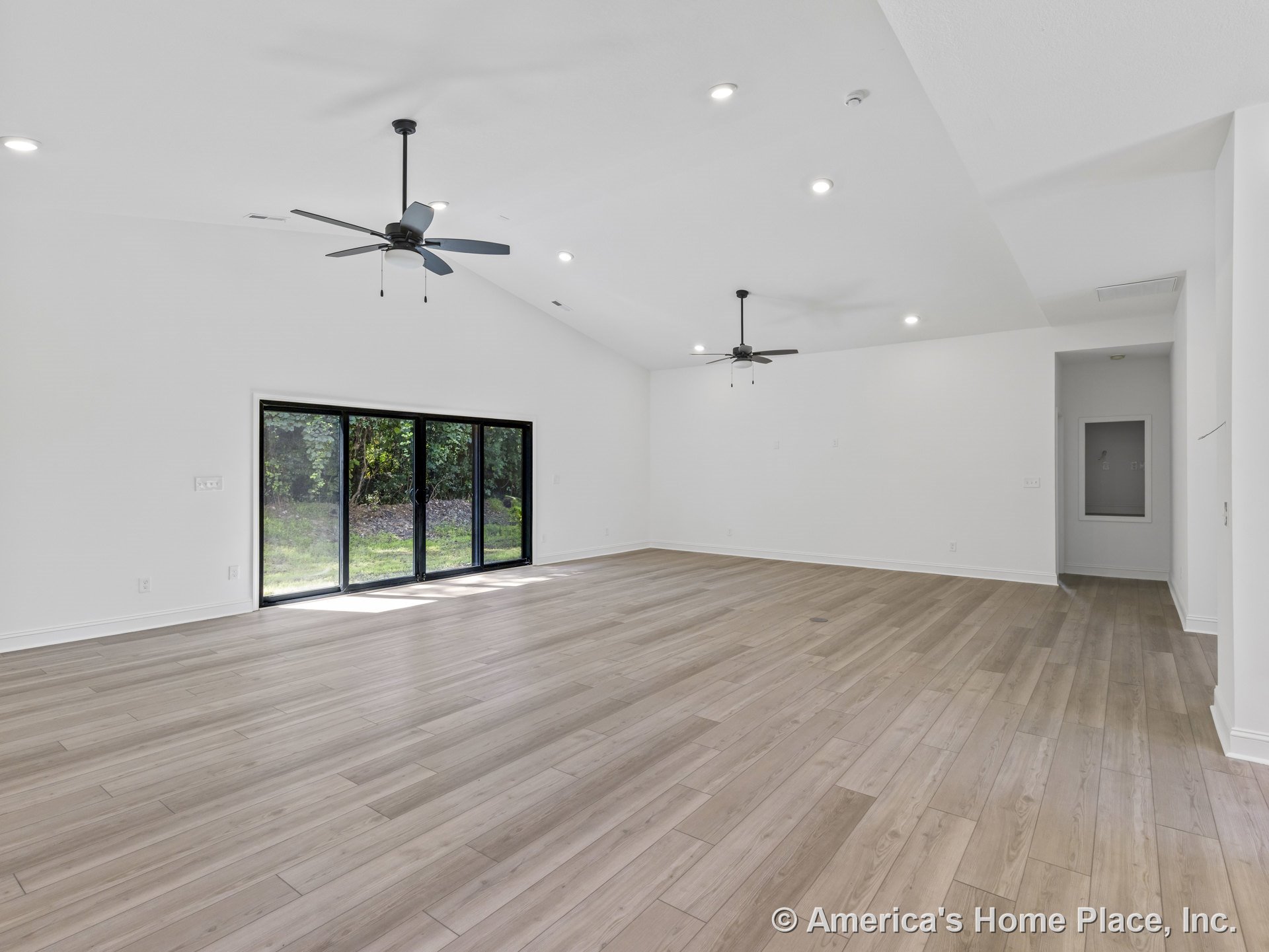 Spacious living room with vaulted ceiling, dual ceiling fans, recessed lighting, light wood plank flooring, large sliding glass doors, white walls and trim, and open floor plan.