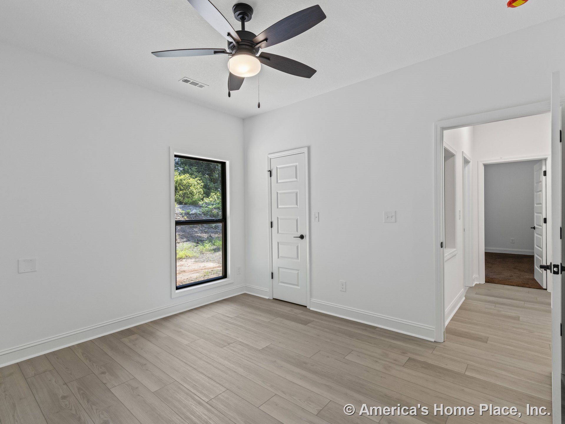 Bedroom with light wood plank flooring, black-framed window, white paneled doors, ceiling fan with integrated light, smooth white ceiling, and white trim and baseboards.