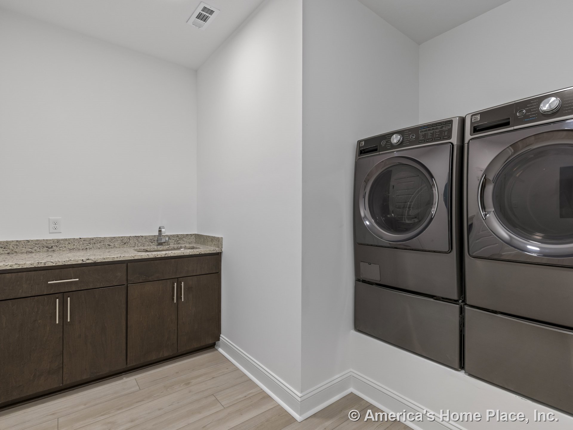Stacked front-loading washer and dryer beside wood cabinets with metal handles, granite countertop with integrated sink, light wood flooring, white walls and ceiling, built-in