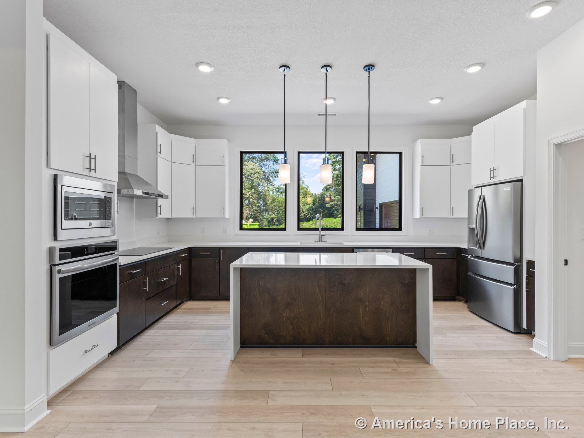 Large kitchen with white upper cabinets and dark lower cabinets, central quartz island, stainless steel range hood, pendant lights above island, light wood flooring, built-in oven