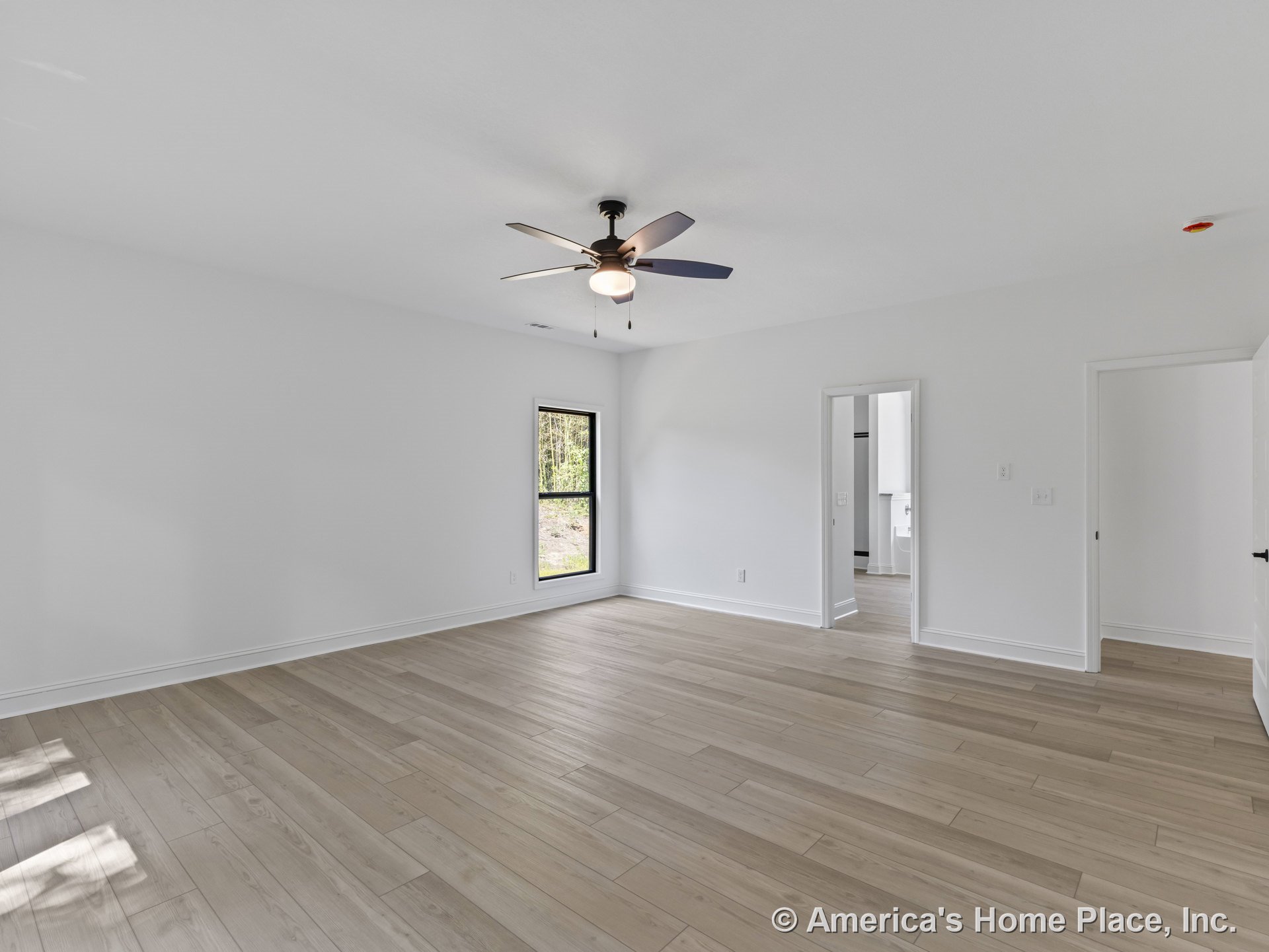 Spacious living room with light wood plank flooring, white walls and trim, black framed window, ceiling fan with integrated lighting, open doorway leading to bathroom, and flush