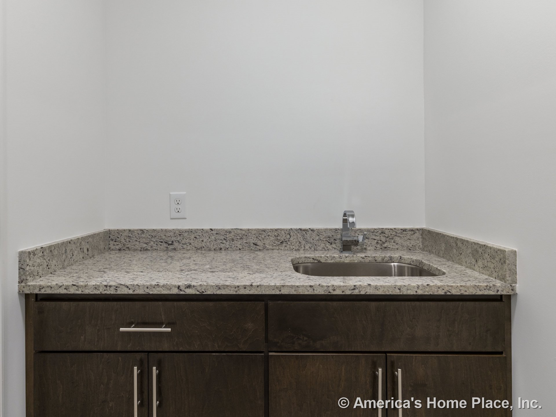 Granite countertop with stainless steel utility sink, modern faucet, dark wood cabinetry, white wall, and built-in electrical outlet.