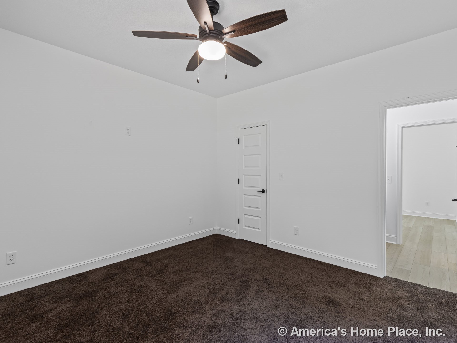 Bedroom with white walls, brown carpet flooring, white baseboards, smooth white ceiling, ceiling fan with light, white paneled door, and visible transition to adjacent room with