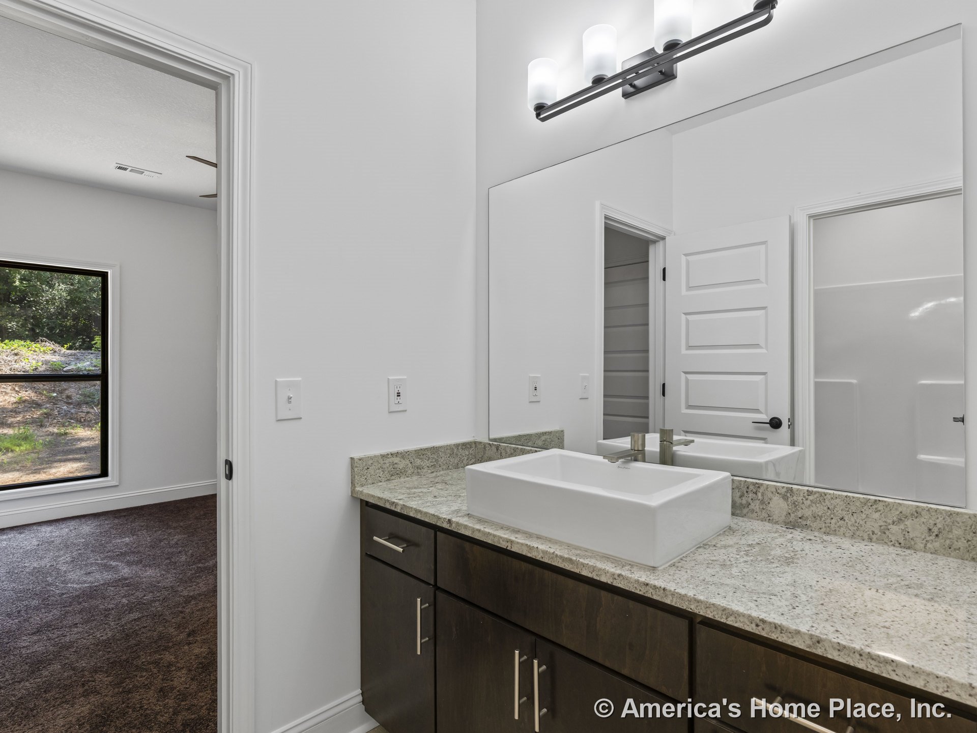 Rectangular vessel sink sits atop a granite countertop with dark wood cabinetry, large wall mirror, modern vanity light fixture, and white paneled door against white walls and
