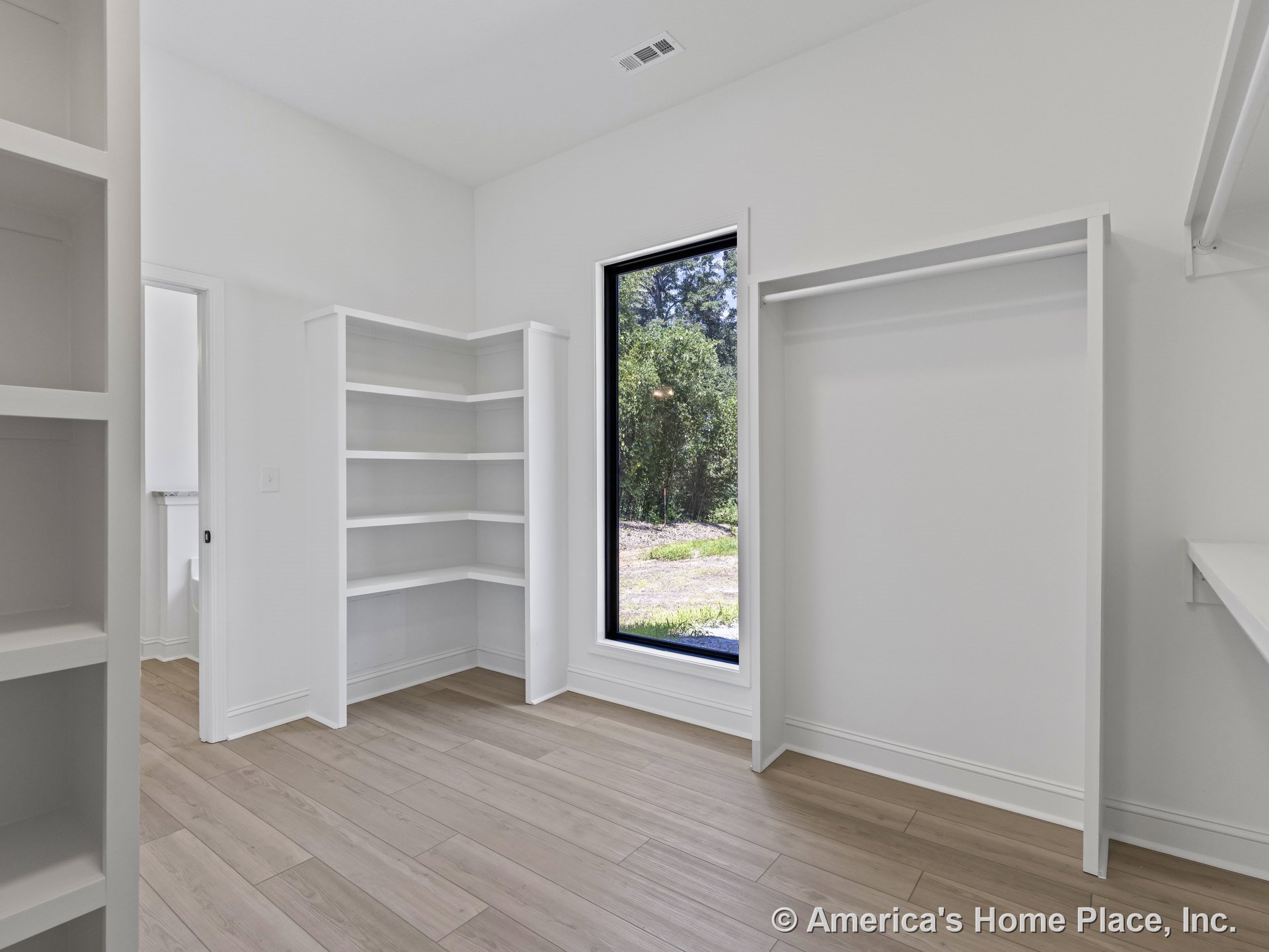 Walk-in closet with built-in corner shelves, open hanging rods, large black-trim window, light wood plank flooring, white walls and trim, and entry to adjacent bathroom.