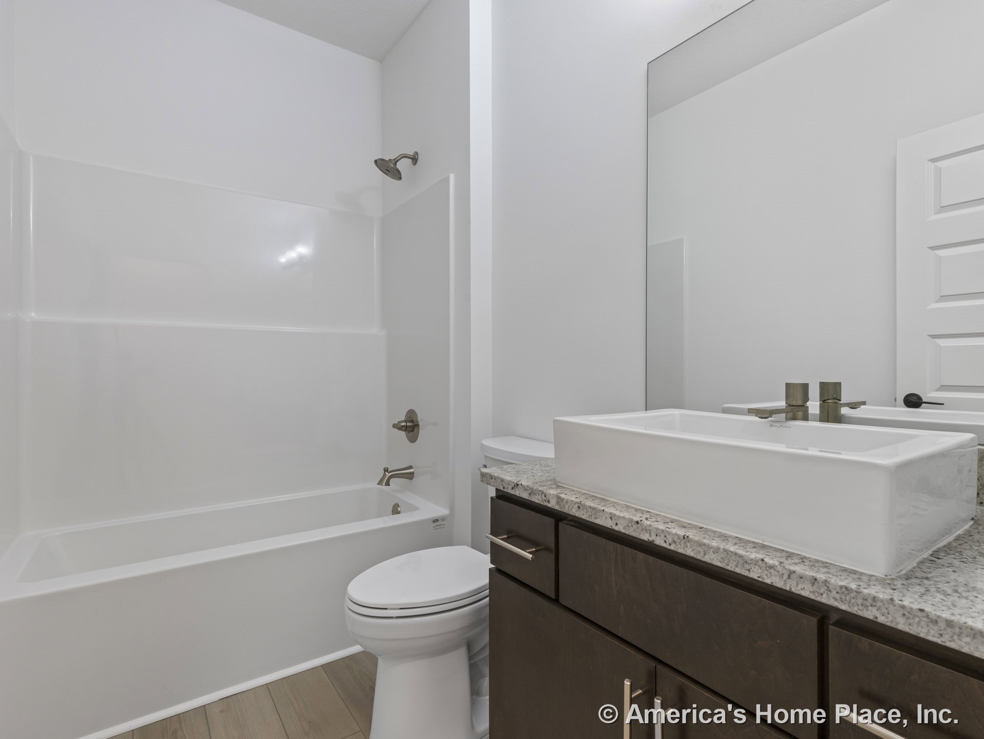 Rectangular vessel sink atop granite countertop with dark wood vanity cabinets, large wall mirror, white bathtub and shower surround, modern faucet, light wood plank flooring