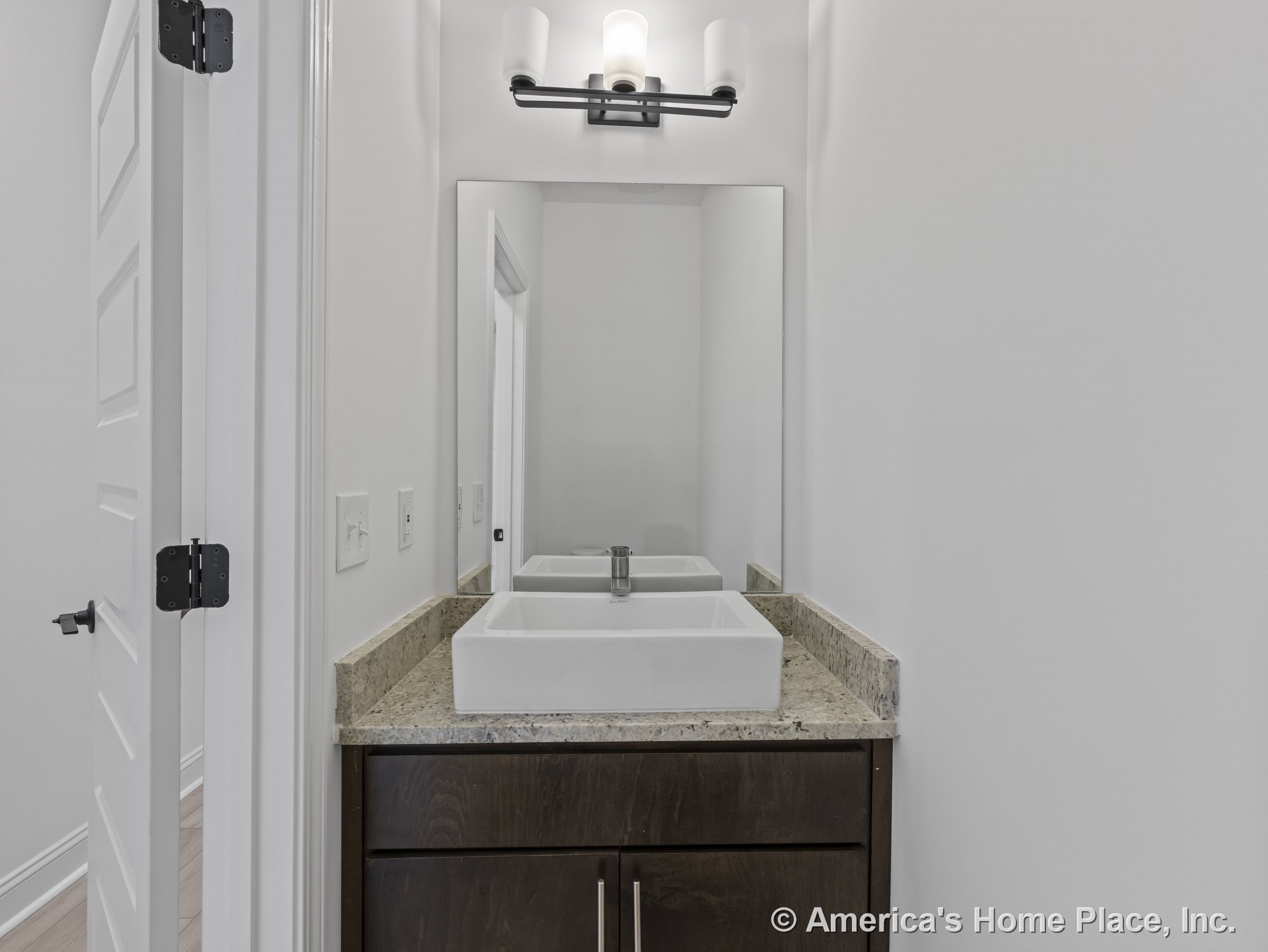 Rectangular vessel sink sits atop a stone countertop with dark wood vanity, large wall mirror, modern wall sconce, and white paneled door surrounded by white walls and door trim.