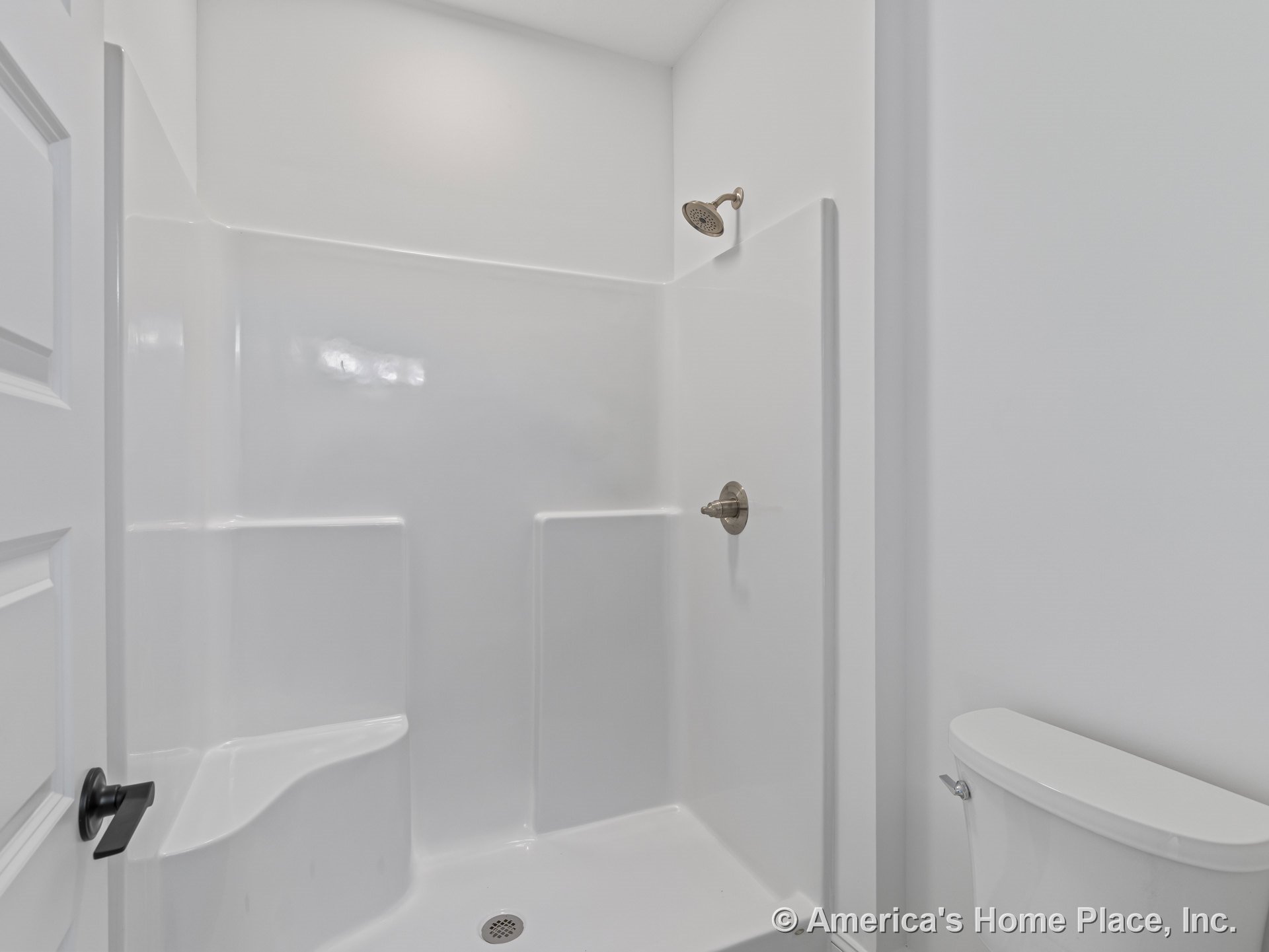 Fiberglass shower enclosure with integrated shelves and single showerhead, adjacent white toilet, white walls, and paneled door featuring black hardware for a minimalist bathroom