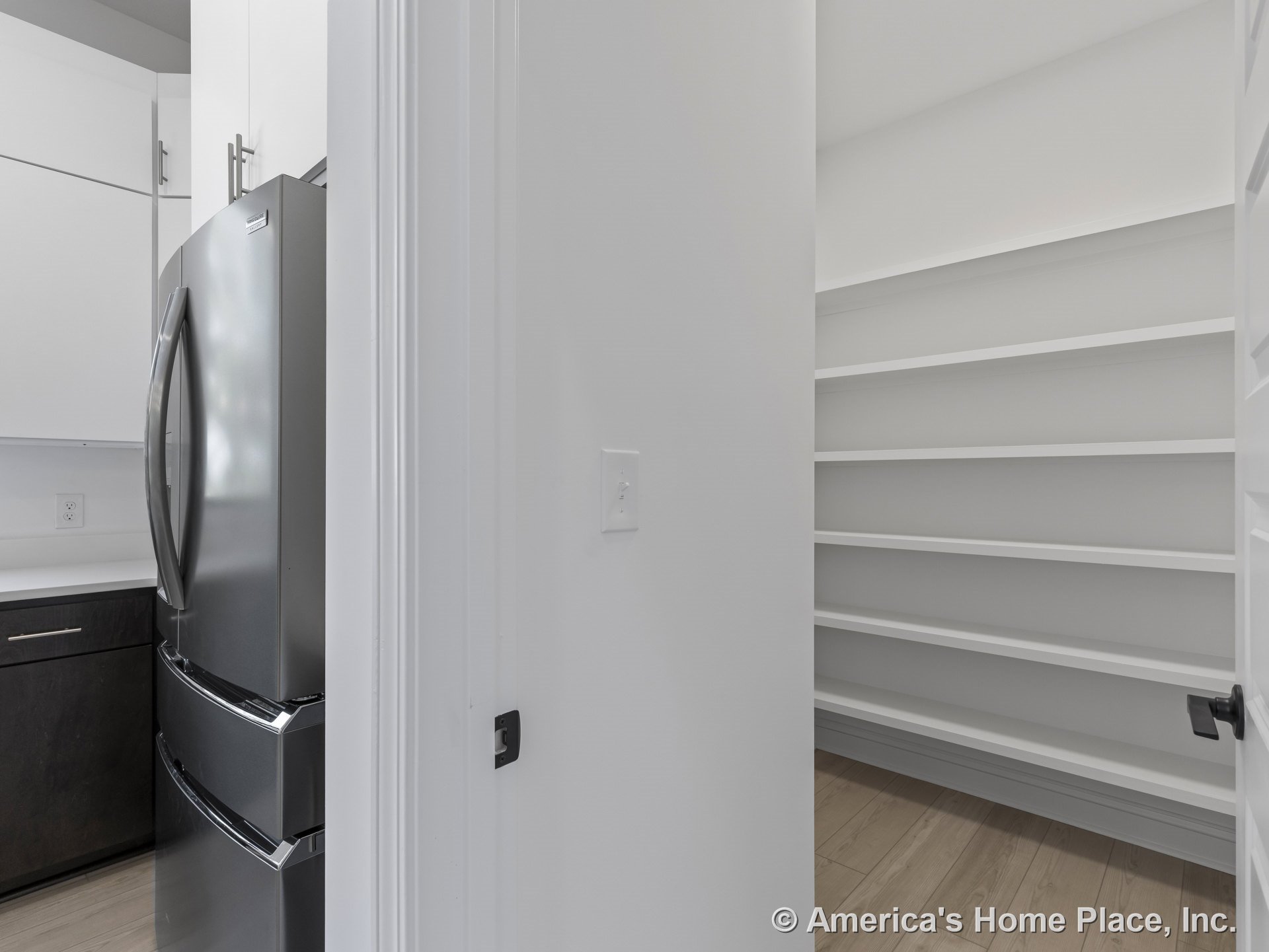 Walk-in pantry with built-in white shelving next to kitchen area featuring dark lower cabinets, stainless steel refrigerator, light wood flooring, and modern finishes.