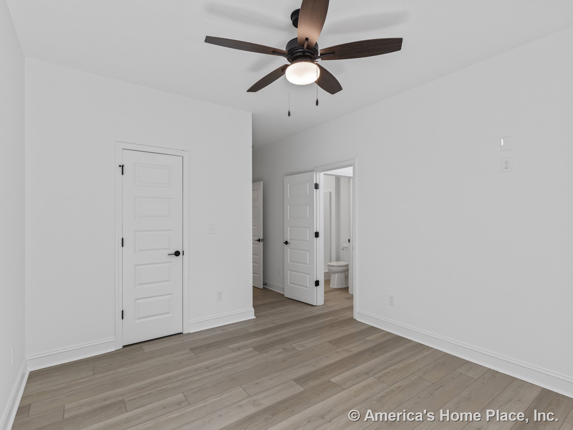 Bedroom with light wood plank flooring, white paneled doors, ceiling fan with integrated light fixture, white baseboard trim, neutral wall color, and visible bathroom entry.