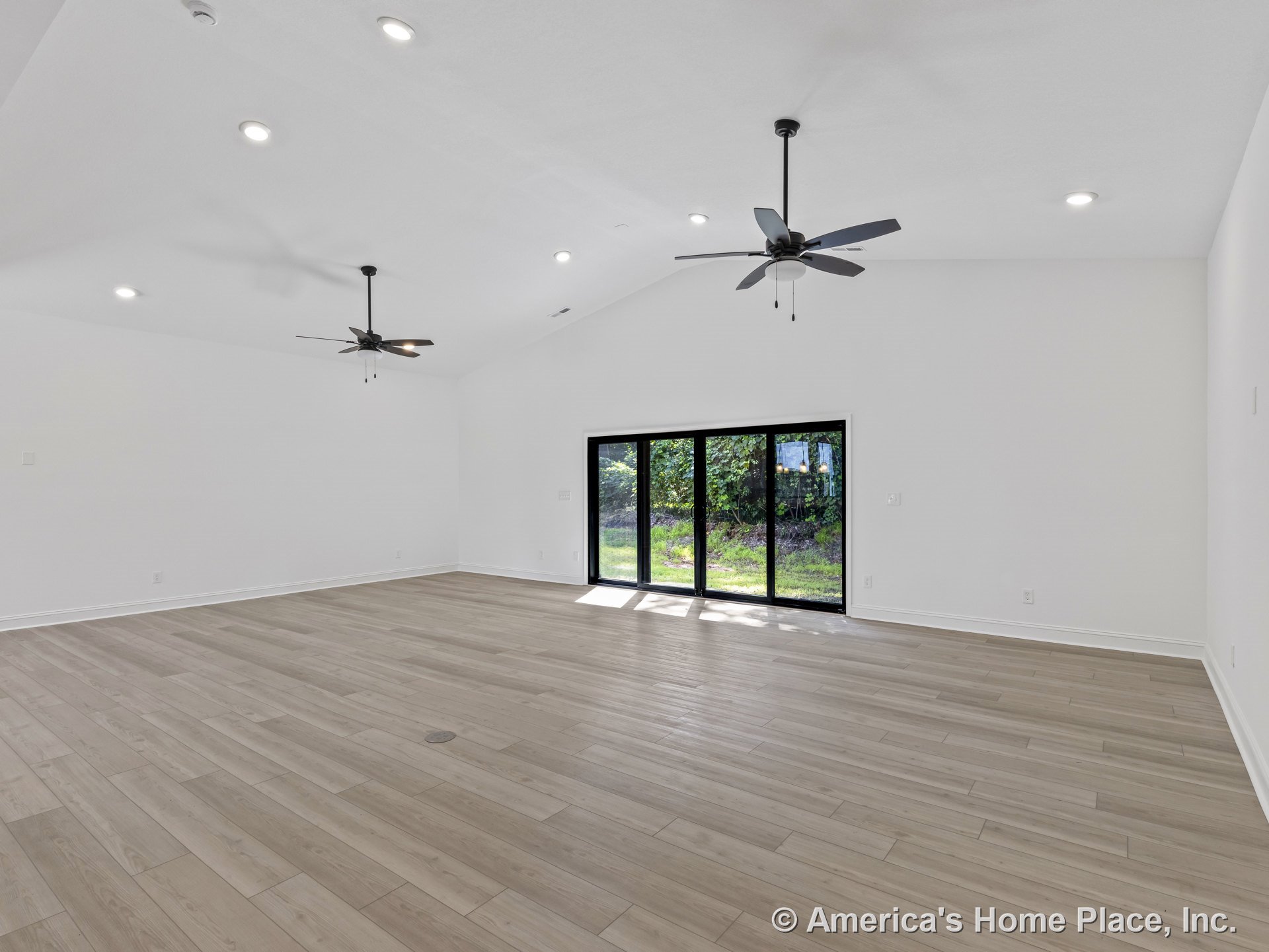 Spacious living room with vaulted ceiling, dual ceiling fans, recessed lighting, light wood plank flooring, large sliding glass doors, white wall trim, and abundant natural light.