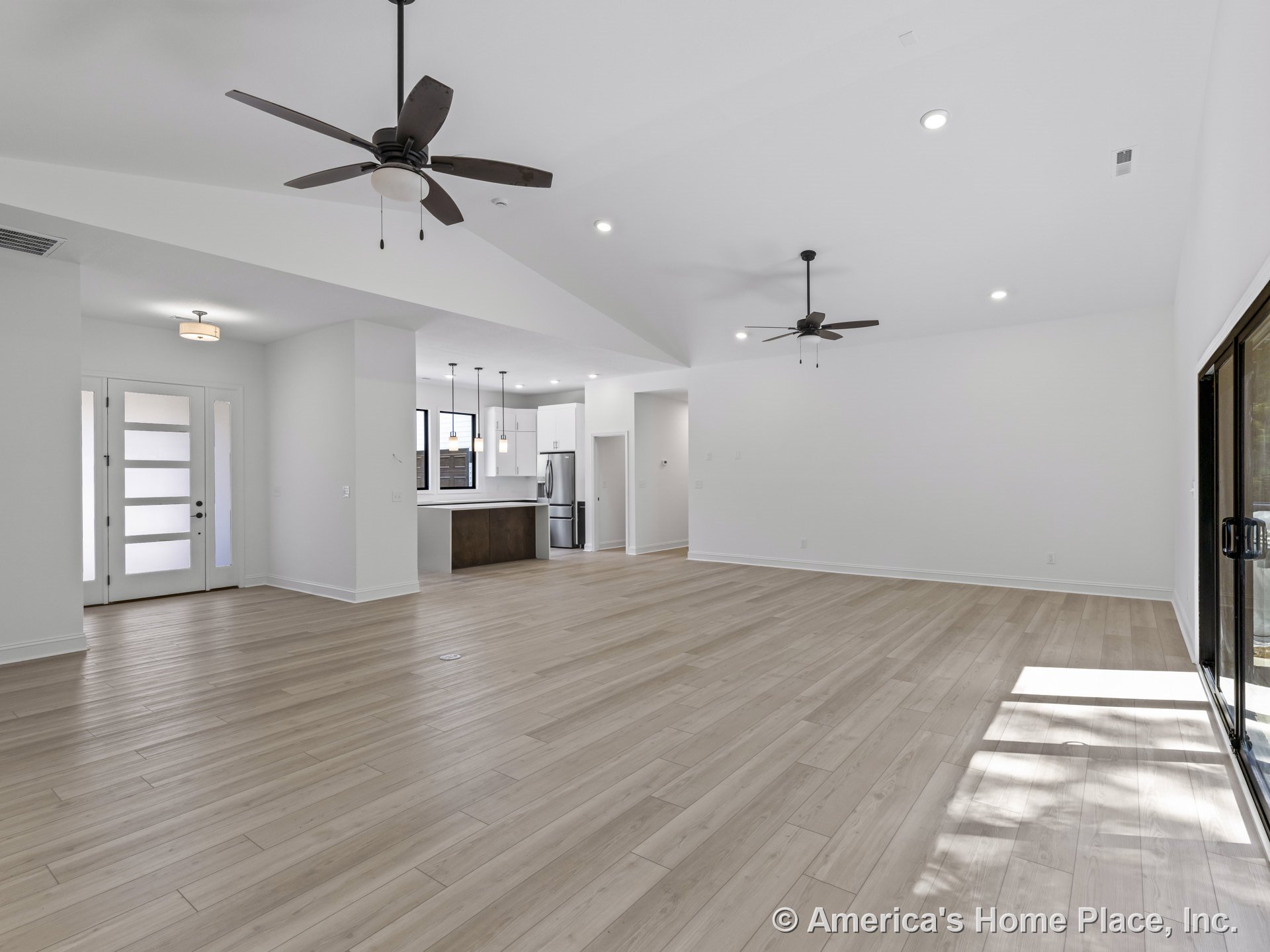 Open-concept living area with light wood plank flooring, vaulted white ceilings, modern ceiling fans, recessed lighting, sliding glass doors, and a contemporary front door.