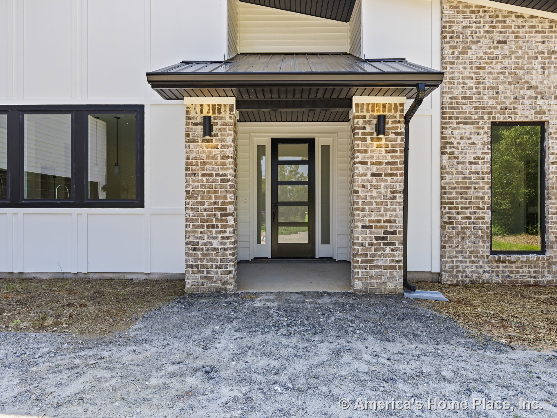 Modern glass front door set beneath a covered porch with brick columns, black framed windows, wall-mounted exterior lights, and white siding with contrasting trim.