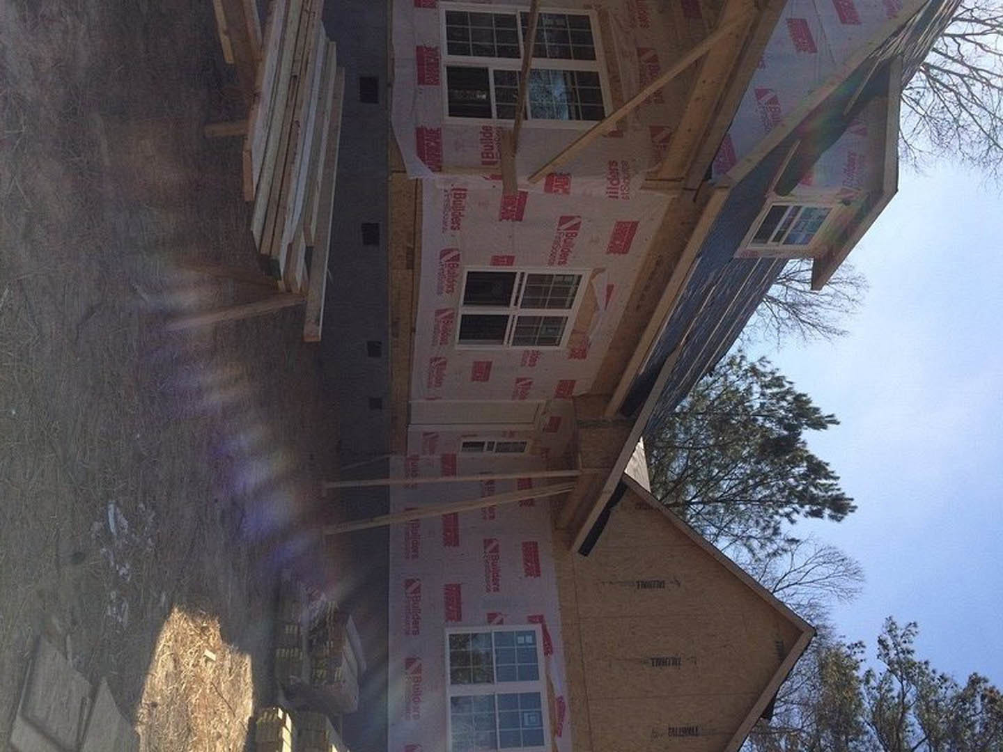 Two-story house under construction with exposed wood planks, partially installed windows, white insulation panels, and black roof tiles; red rectangular object sits on a white