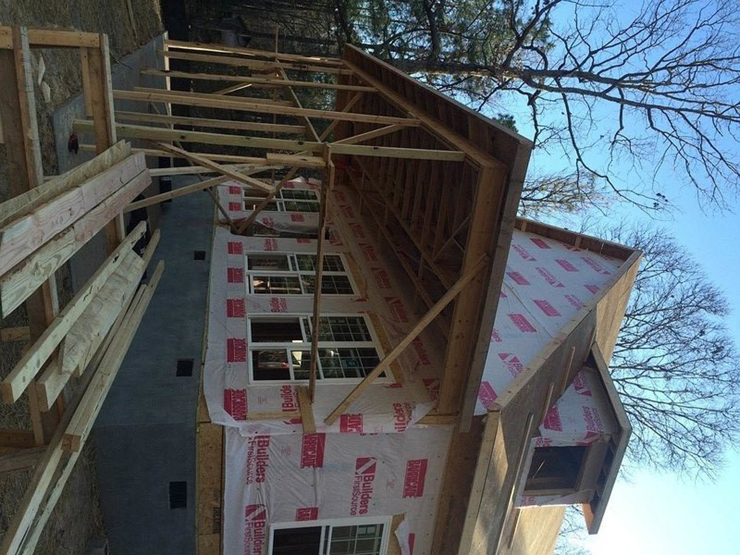 Wood-framed house under construction with exposed windows, ladder leaning against exterior, red and white sign near entrance, unfinished roofing, surrounded by trees and sky.