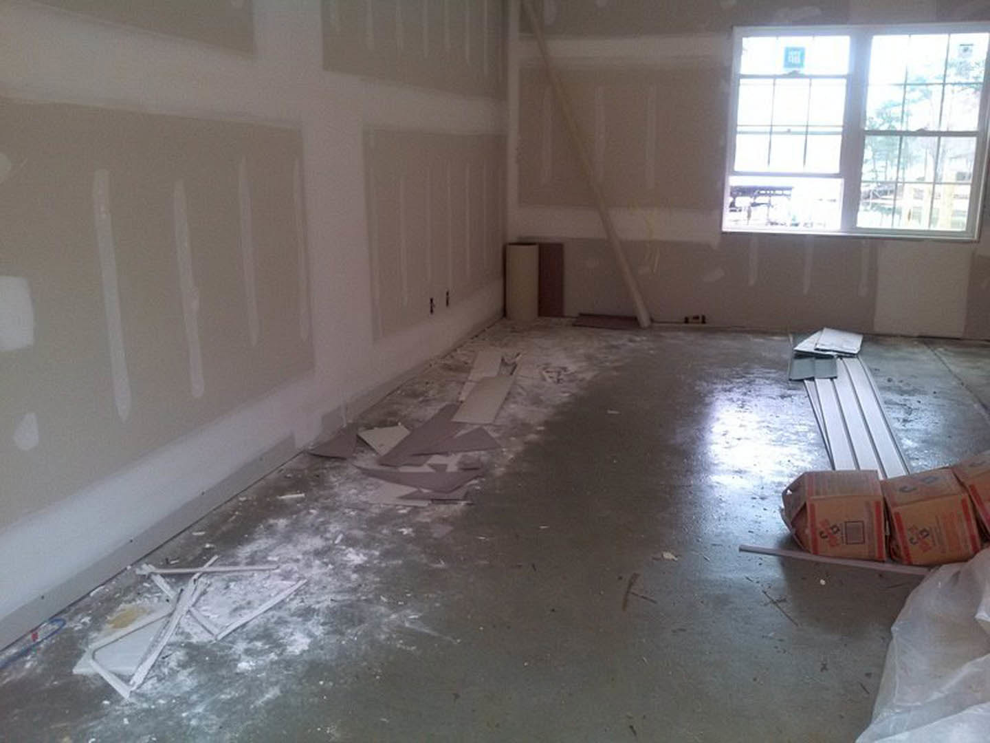 White plaster wall with brown window frame, blue sign taped to window, two cardboard boxes on sill, broken white object and debris scattered on composite flooring