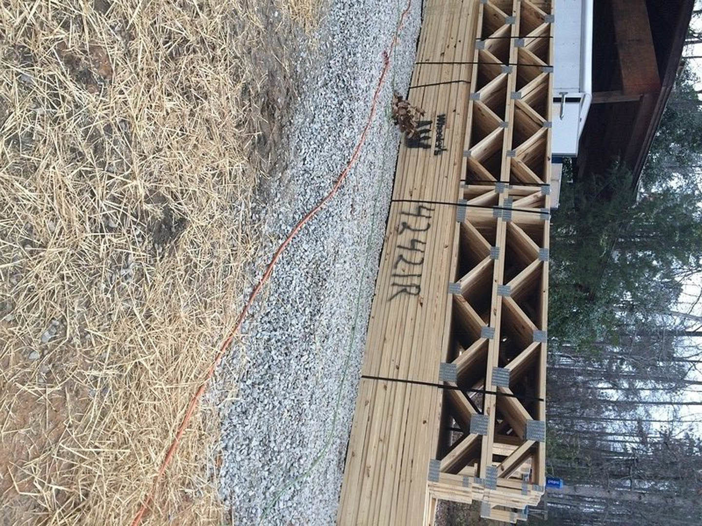 Stack of wooden trusses and lumber on dry grass, red rope nearby, wooden box with holes and writing, trees in background.