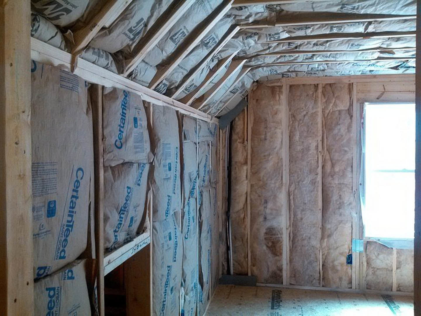 Room under construction with exposed wood framing, insulated walls and ceiling, window letting in natural light, white electrical box with blue text, and unfinished white floor.