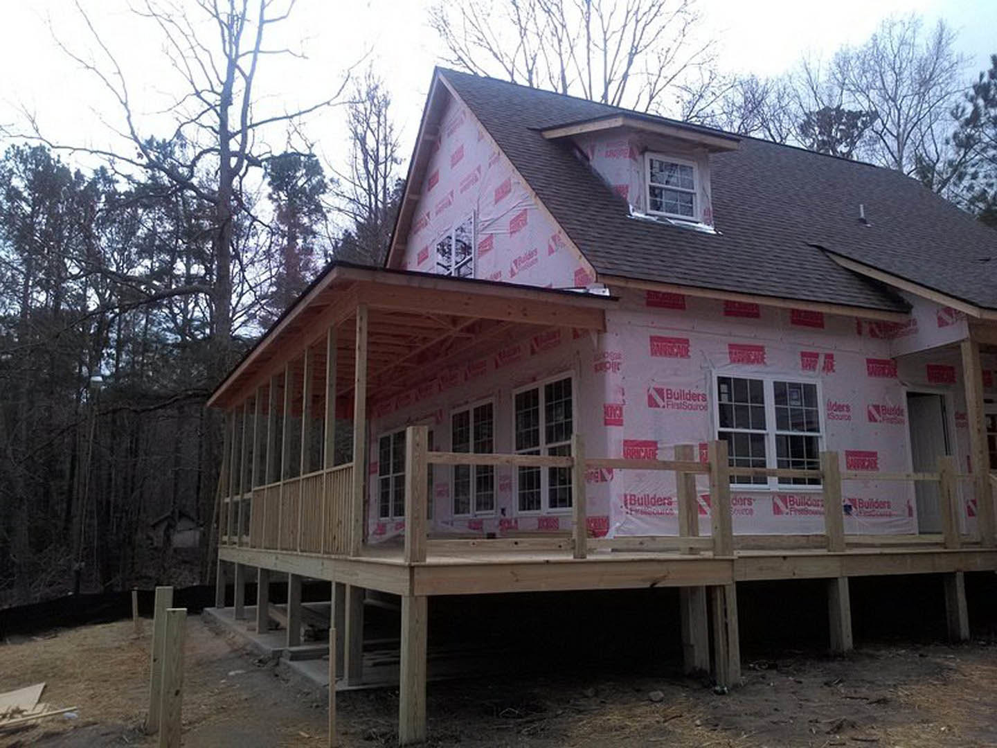 Partially built house with exposed lumber framing, red tape sealing window edges, unfinished siding, and construction materials scattered around exterior porch area